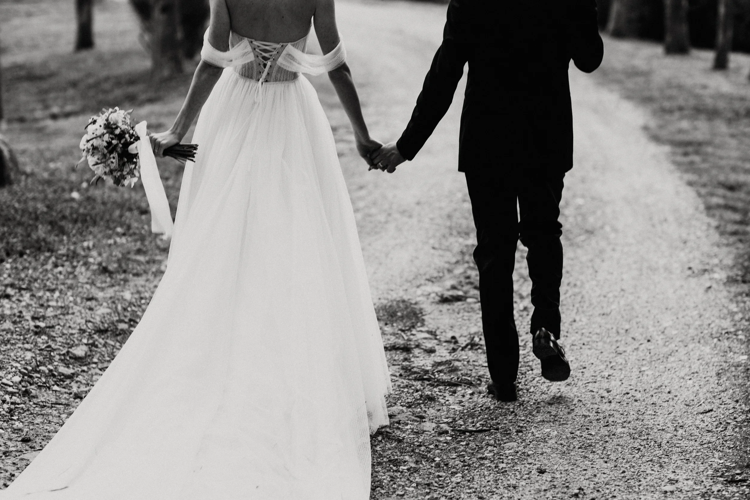 Una coppia di sposi che cammina mano nella mano su un sentiero, l'uomo indossa un abito nero e la donna un vestito da sposa bianco, vista da dietro in bianco e nero. Villa dal Pozzo