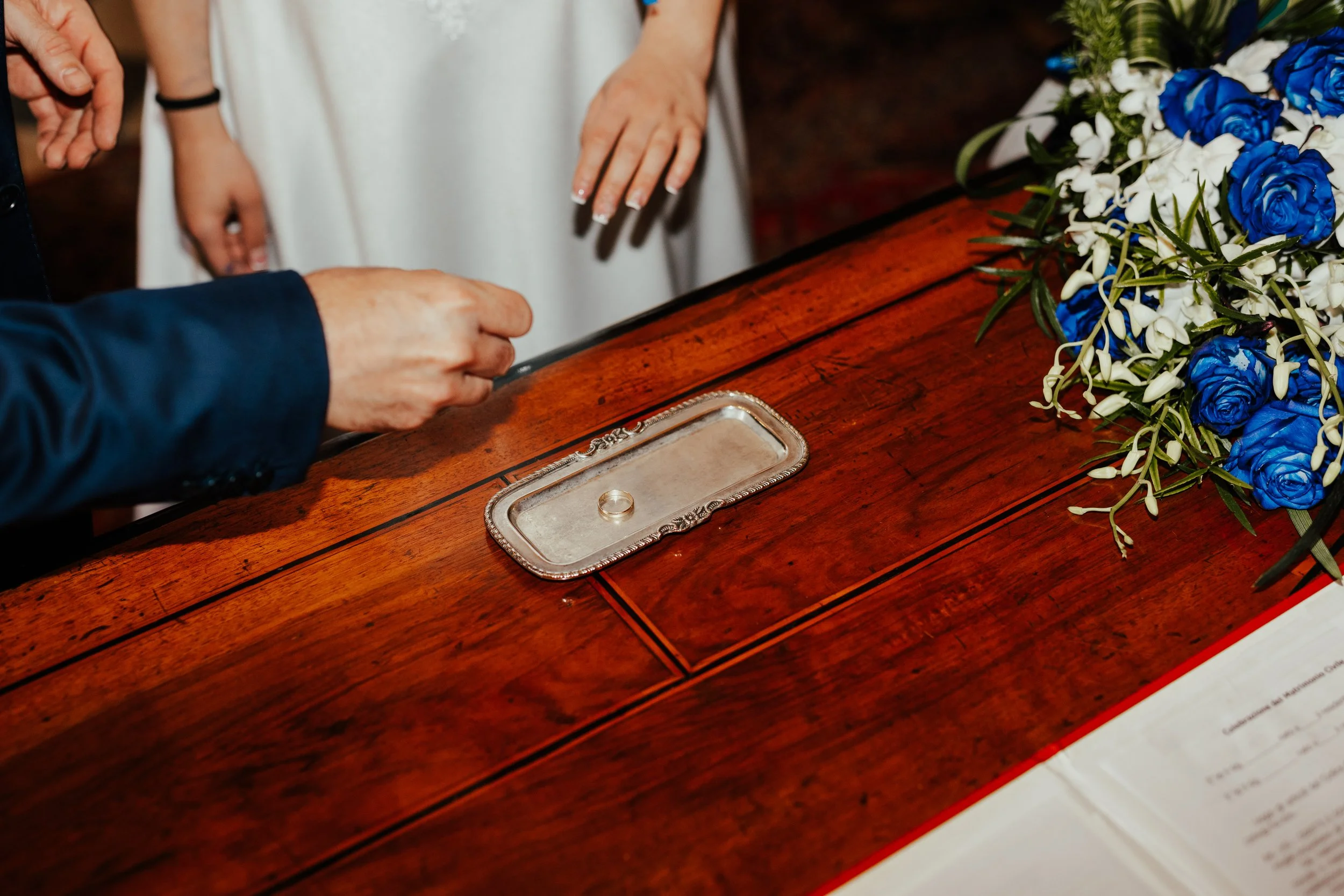 Un'ambientazione di nozze con mani che scambiano l'anello sul tavolo di legno, decorato con mandorli blu e bianchi.