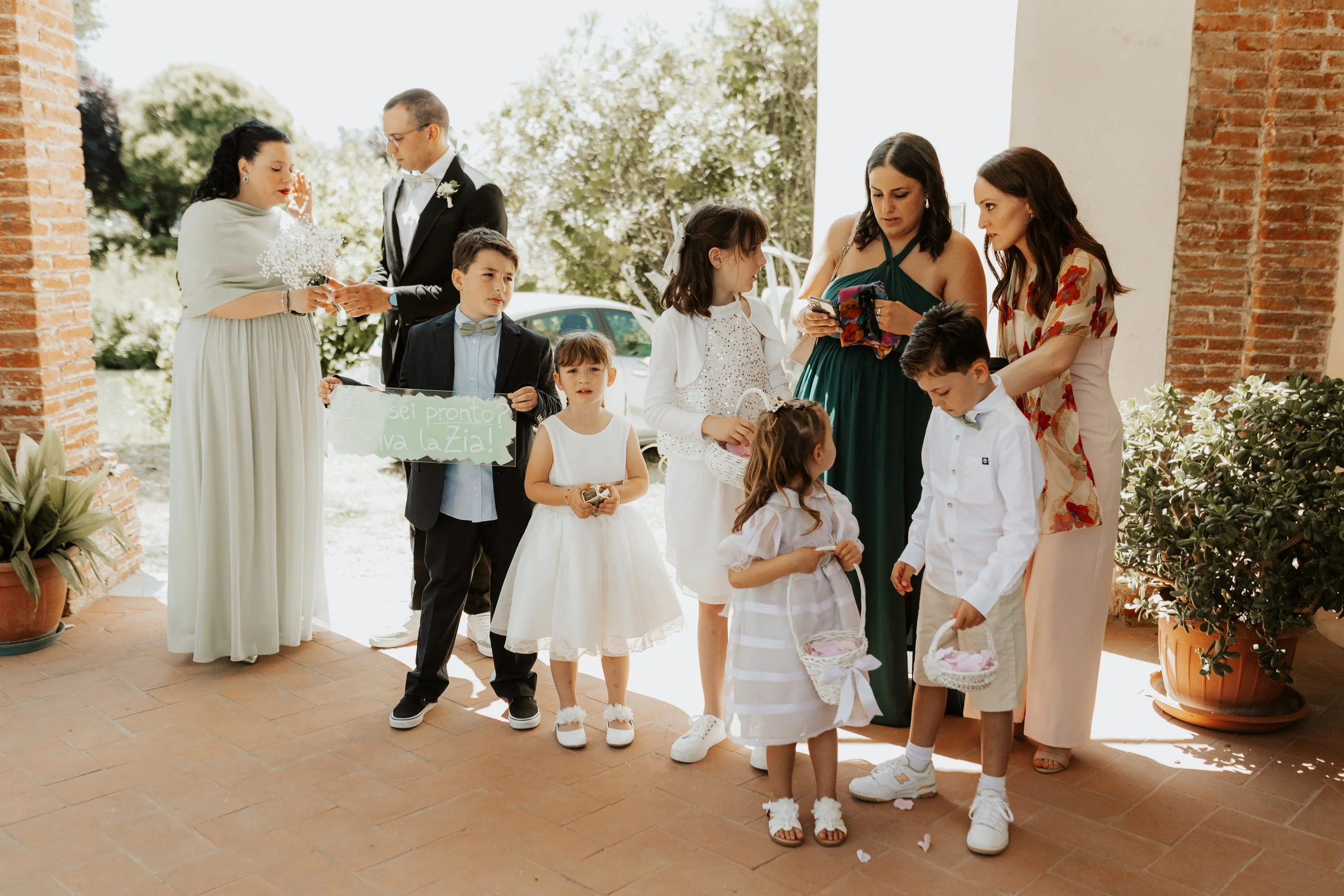 Un gruppo di persone, tra adulti e bambini, sulla terrazza di un edificio in bianco e mattoni, probabilmente in attesa di un matrimonio, con alcuni adulti che osservano i bambini. C'è una donna con abito bianco e un uomo con abito da sposo, e i bambi