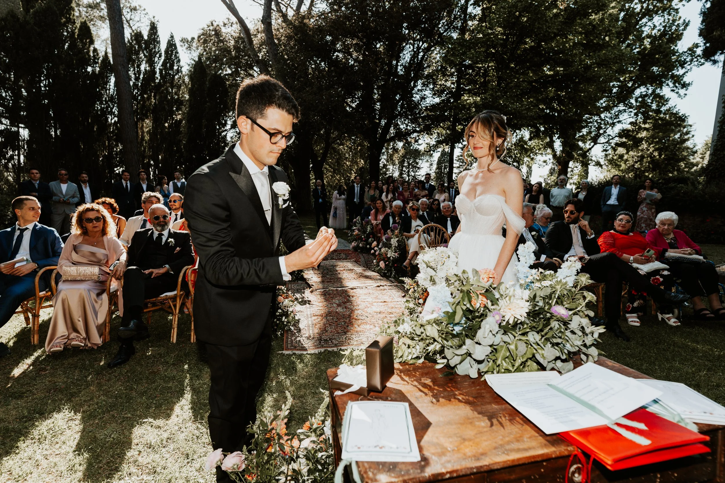 Sposi che si scambiano le promesse di matrimonio durante una cerimonia all'aperto con amici e familiari seduti. La sposa indossa un abito bianco e il sposo un completo nero. Sul tavolo ci sono fiori e documenti.