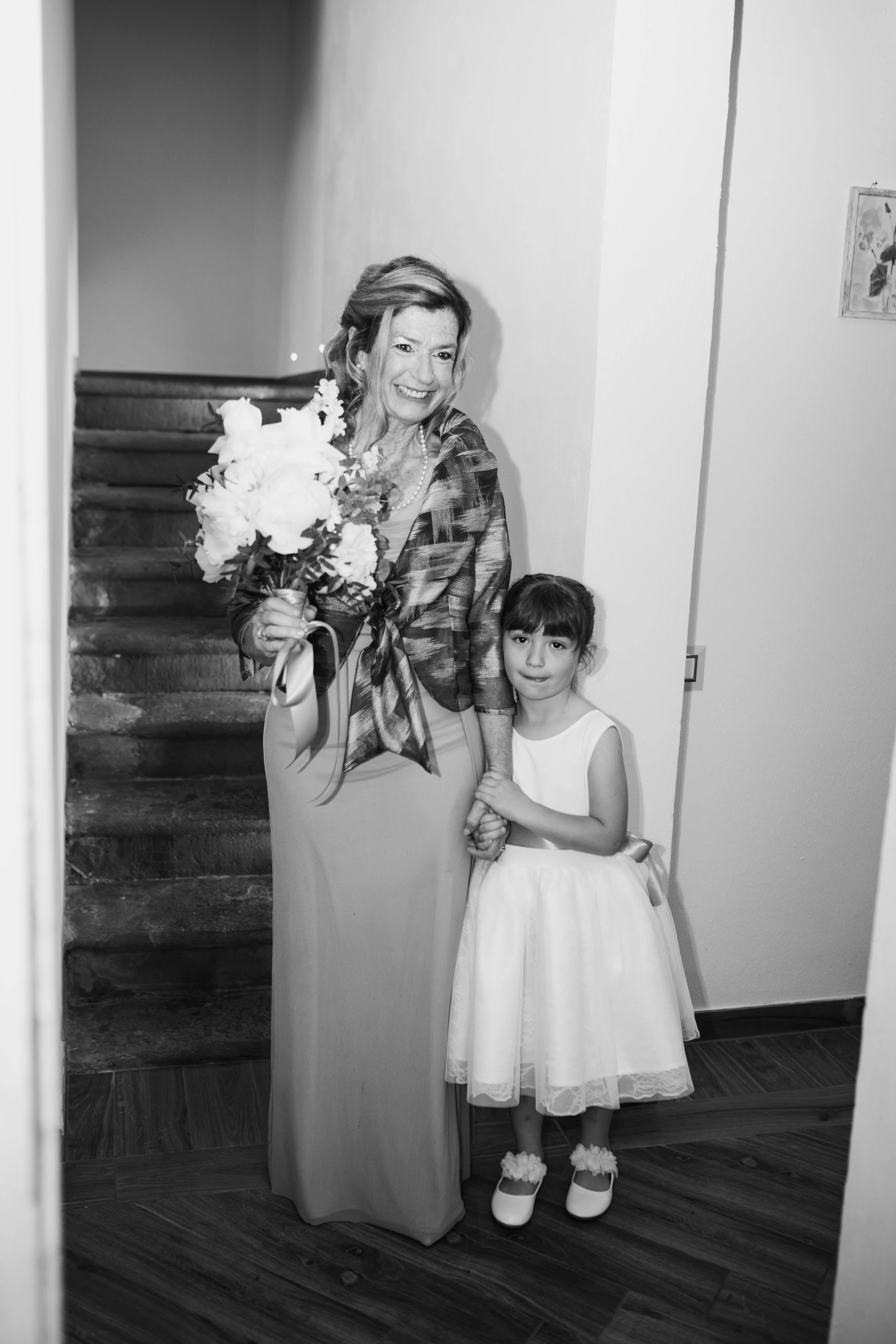 Una donna anziana e una bambina stanno insieme in un corridoio, sorridendo. La donna tiene un mazzo di fiori e indossa un abito lungo con una giacca, mentre la bambina indossa un vestito bianco con scarpe abbellite da fioretti.
