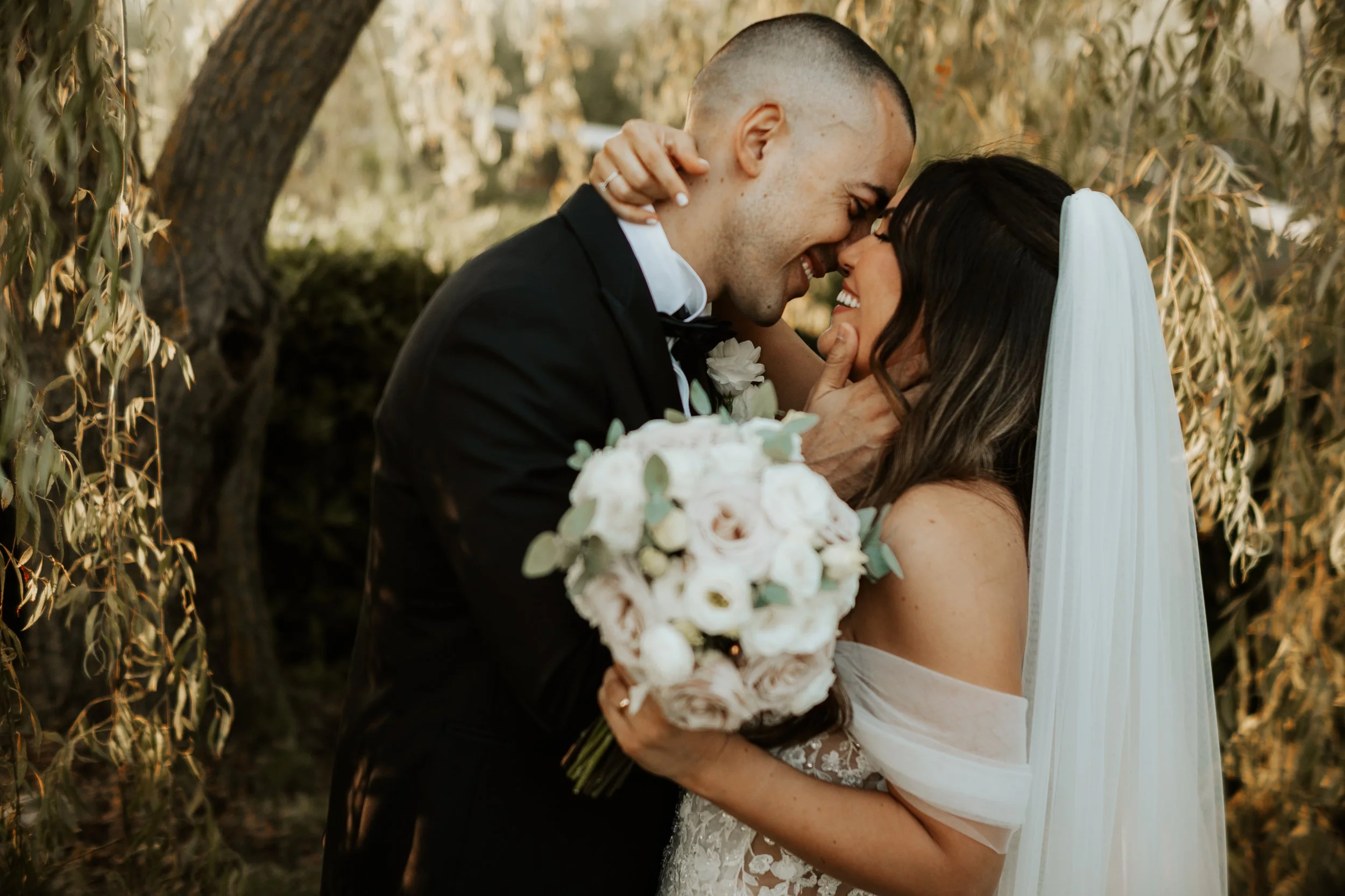 Una coppia di sposi abbracciati, sorridenti e felici, si prepara a baciarsi durante il loro matrimonio all'aperto, con un bouquet di fiori bianchi.
