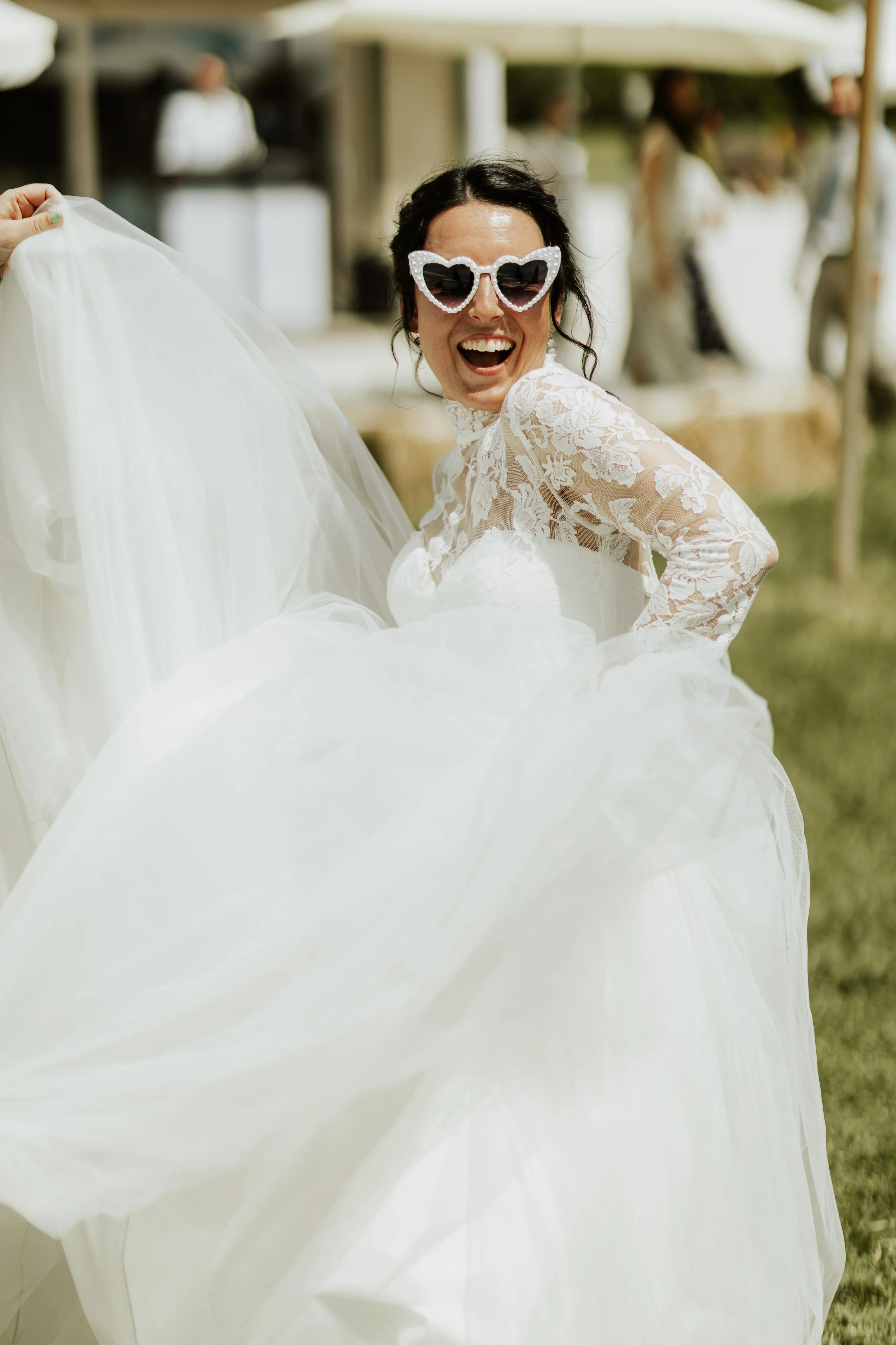 Una donna sorridente con occhiali a cuore bianchi indossa un abito da sposa bianco in un contesto all'aperto.