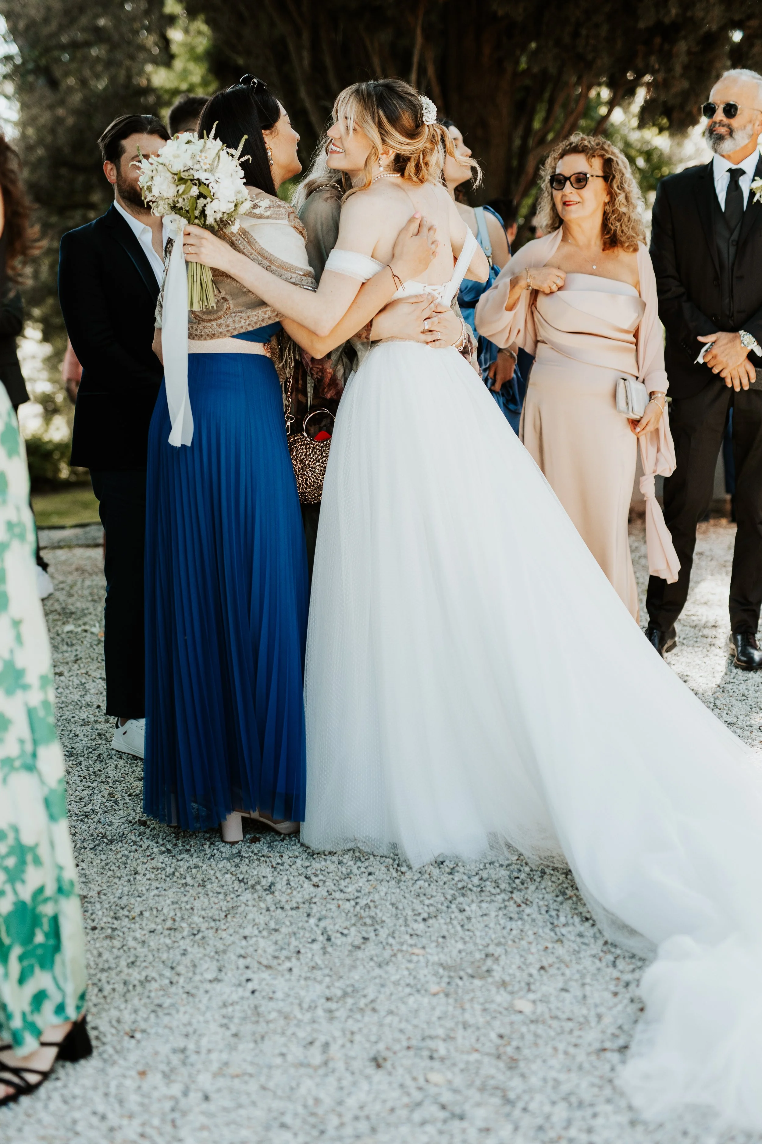 Un gruppo di persone alla cerimonia di matrimonio, con alcune donne che si abbracciano e sorridono, alcune portando abiti eleganti, e un bouquet di fiori.