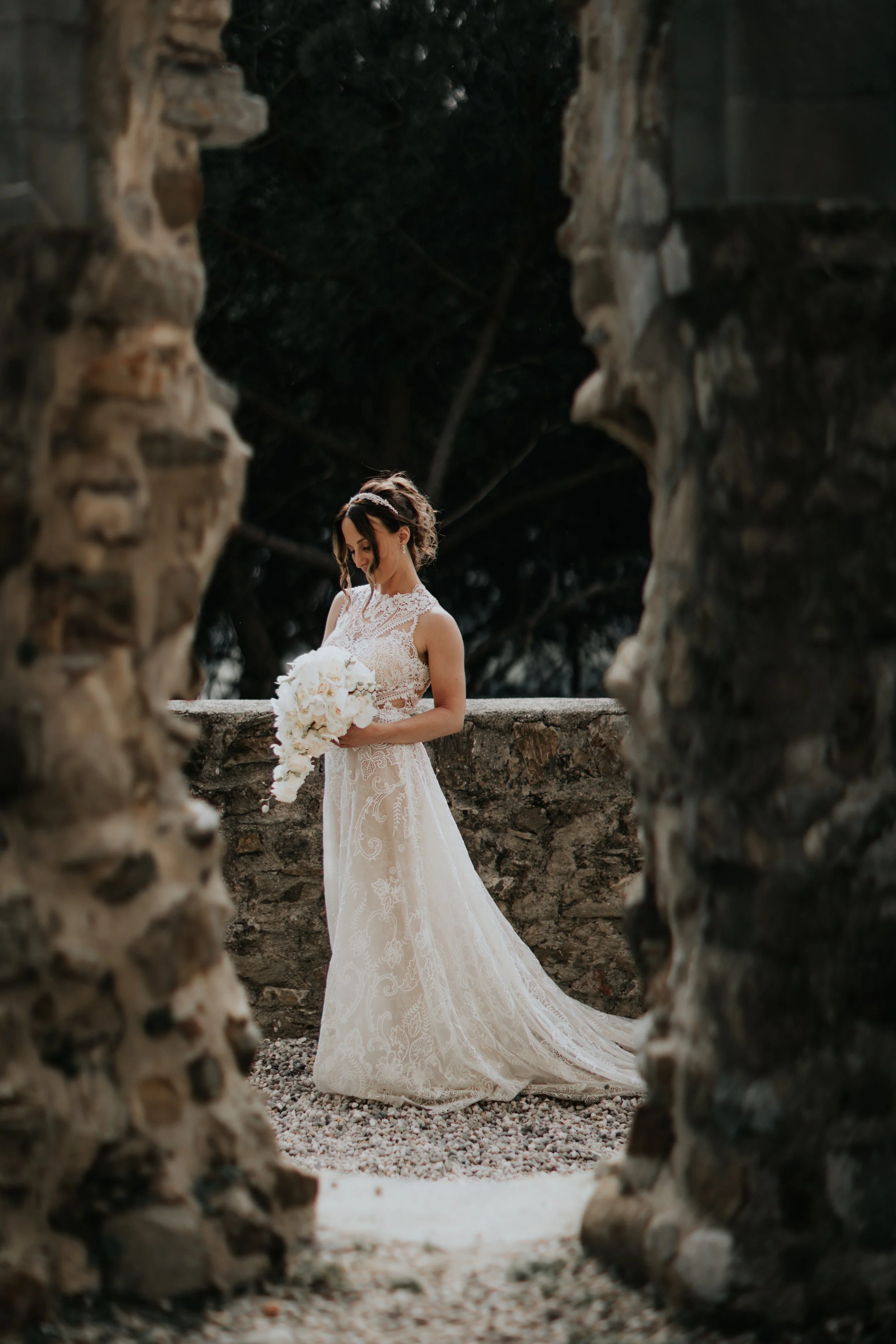 Una donna in abito da sposa bianco con un bouquet di fiori chiari, vista attraverso una cornice di pietre, in un ambiente all'aperto con sfondo scuro e alberi.