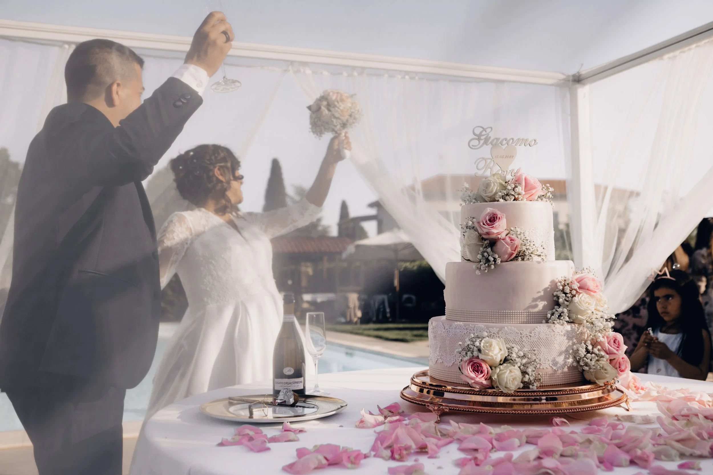 Una coppia di sposi festeggia il matrimonio davanti a una torta decorata con rose, con petali di rosa sparsi sul tavolo, sotto una tensostruttura con tende di garza bianca.