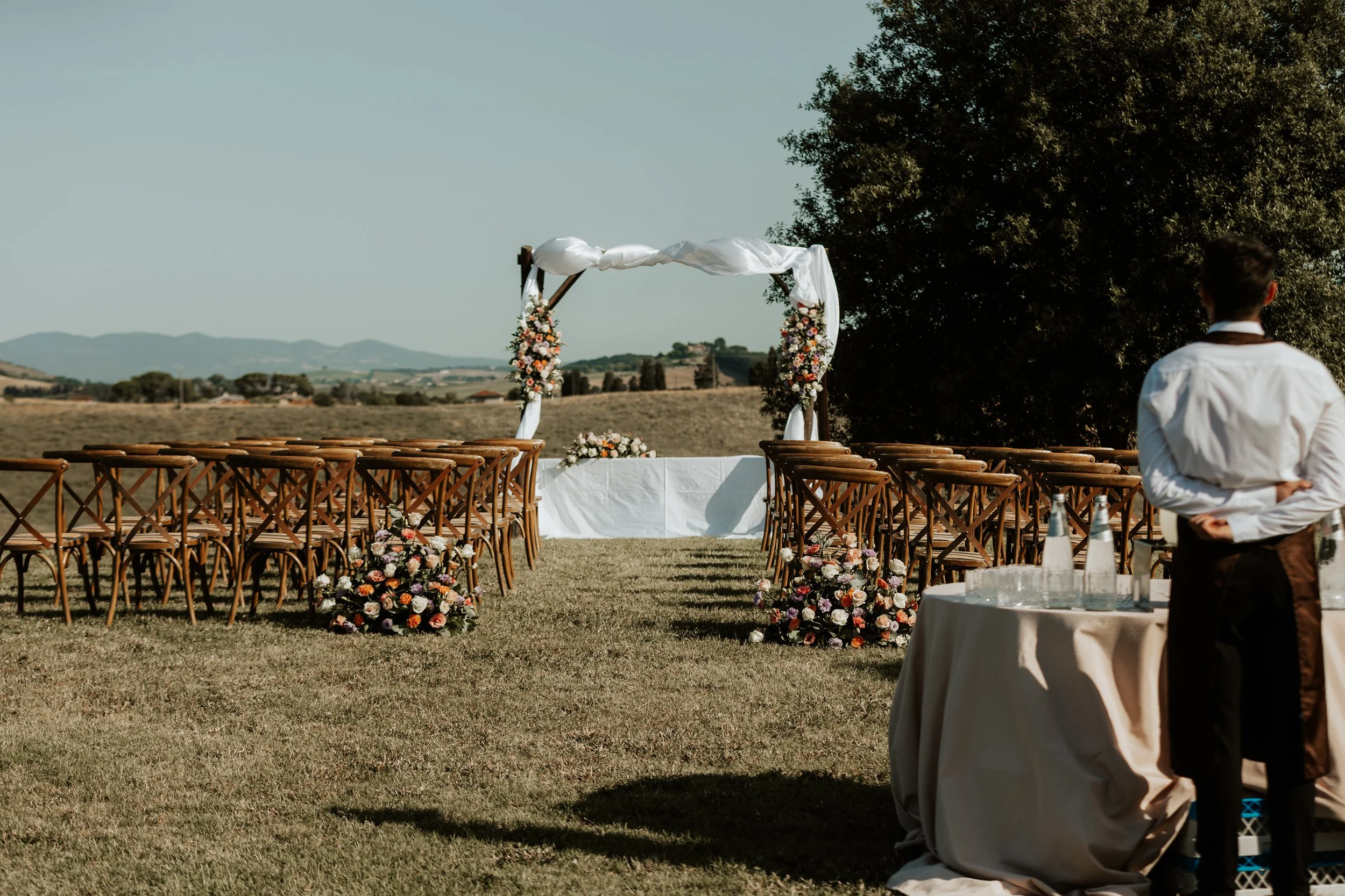 Setup per matrimonio all'aperto con cerimonia sotto un arco decorato con fiori, su un prato con vista sulle colline. Agriturismo Cà Lo Spelli. Del Carlo Catering