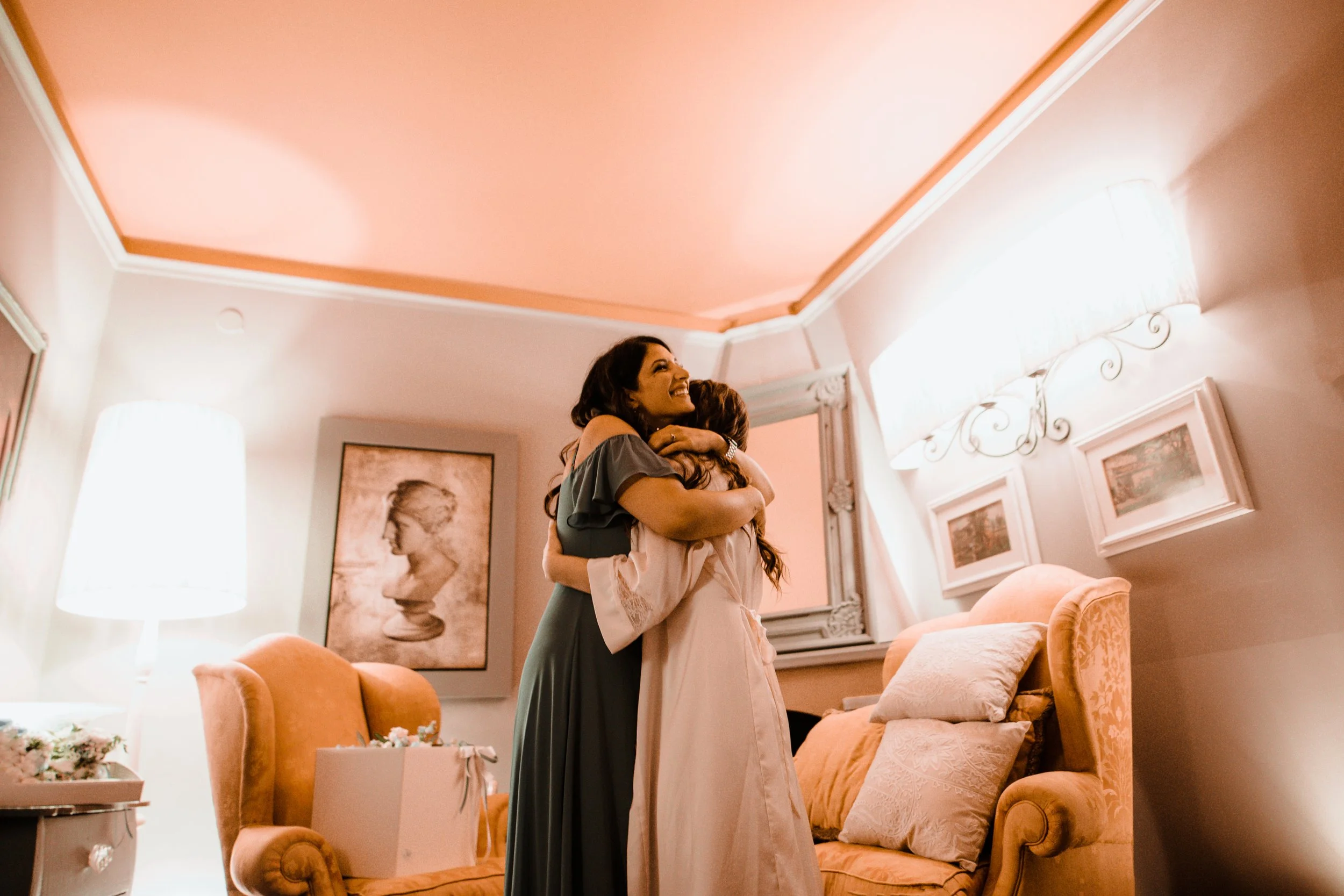 Due donne si abbracciano felici in un soggiorno accogliente con pareti rosa, sedie imbottite, e illuminazione calda.