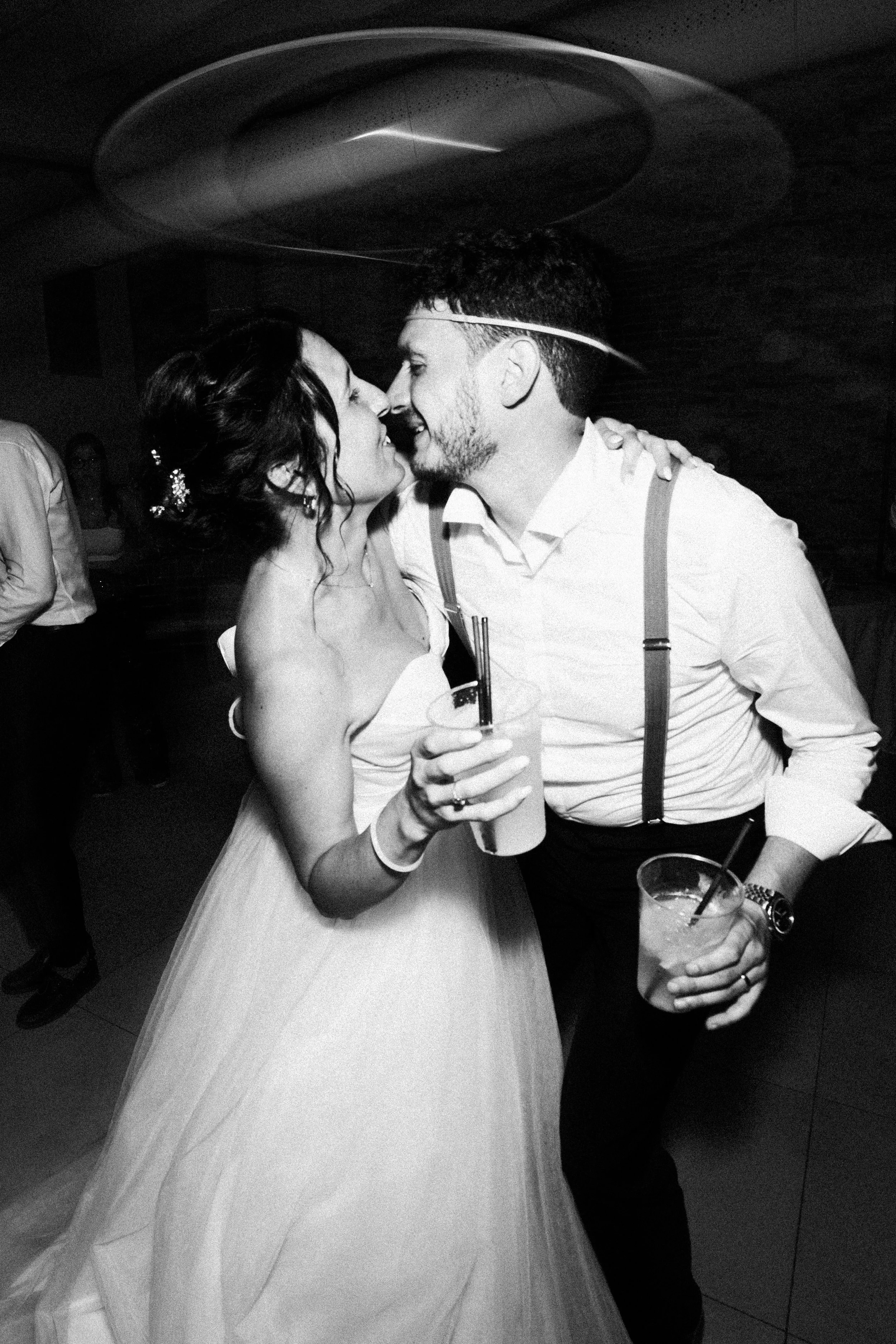 Una coppia di sposi si bacia e sorride durante la loro festa di matrimonio, entrambi con abiti eleganti, mentre tengono delle bevande.
