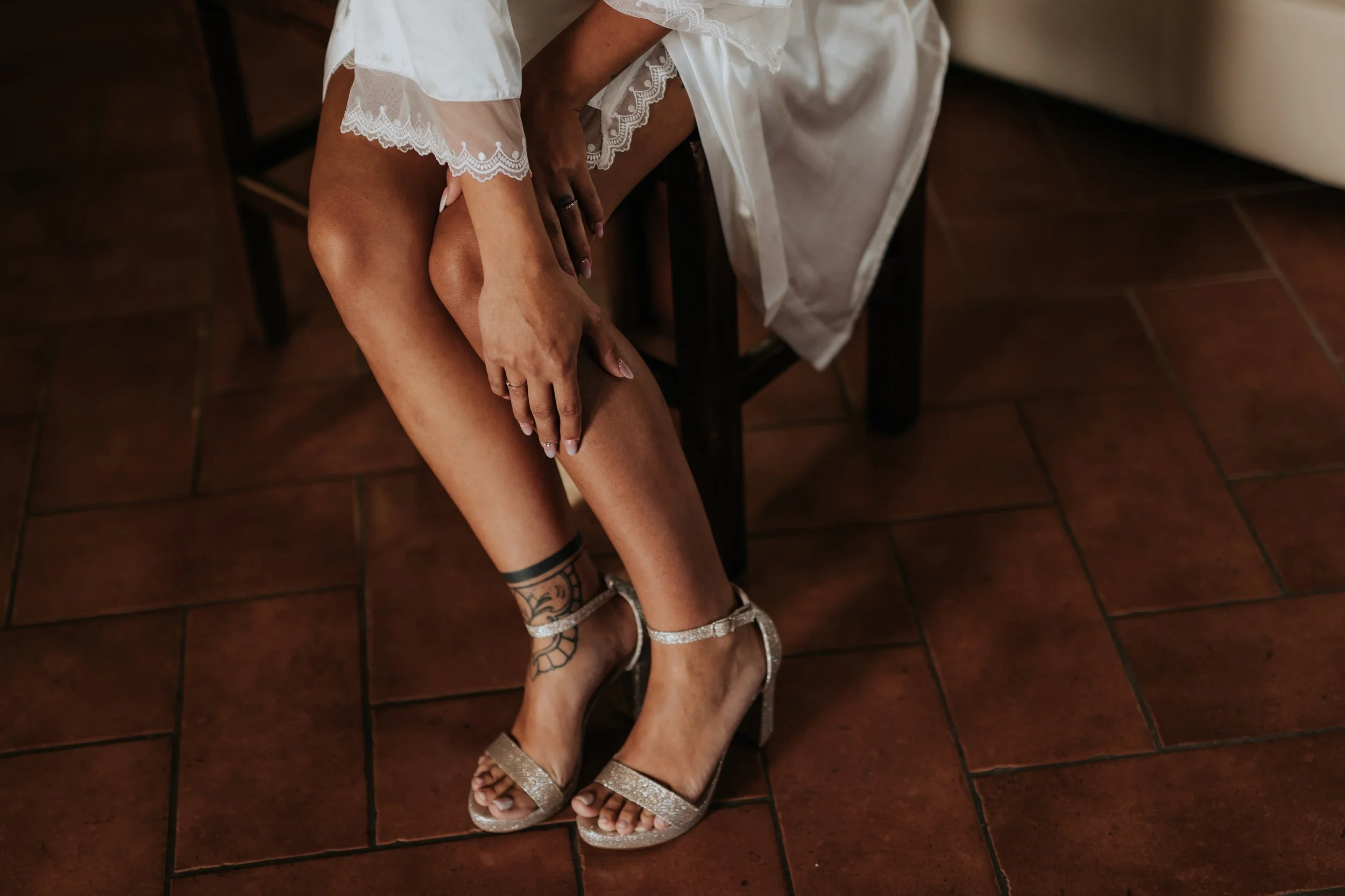 Una persona con scarpe da sera glitterate e tatuaggi sulle gambe si sistema le gambe, seduta su una sedia di legno, con una gonna bianca con pizzo. Il pavimento è di piastrelle rosse.