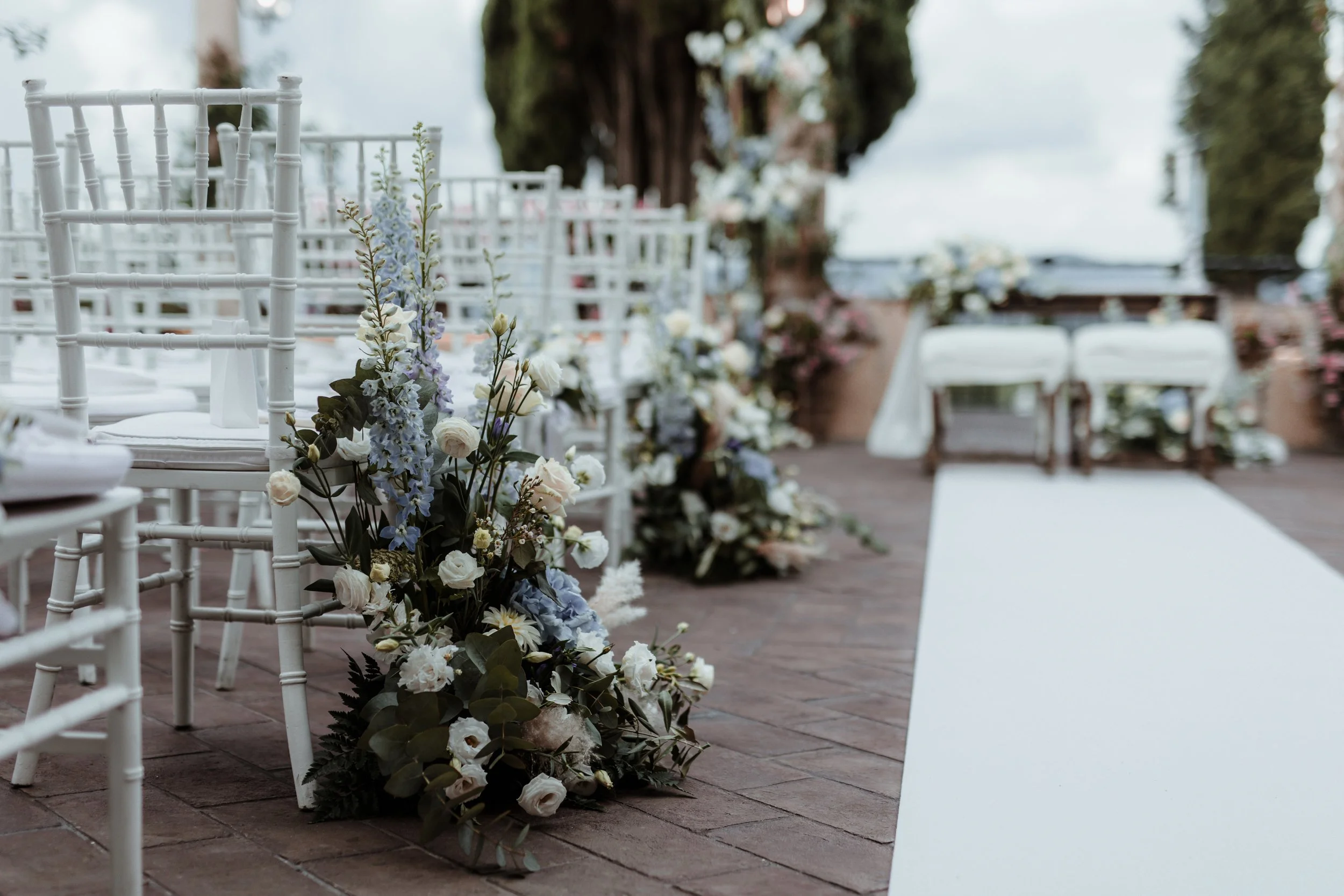 Setup per matrimonio con sedie bianche, fiori e decorazioni floreali lungo il corridoio all'aperto.