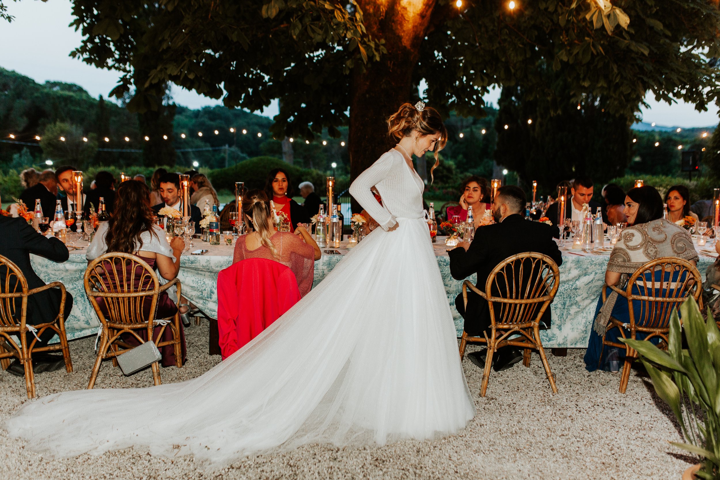 Una sposa con un vestito lungo e un velo bianco si trova vicino a un tavolo decorato per un matrimonio, circondata da invitati seduti in un giardino illuminato.