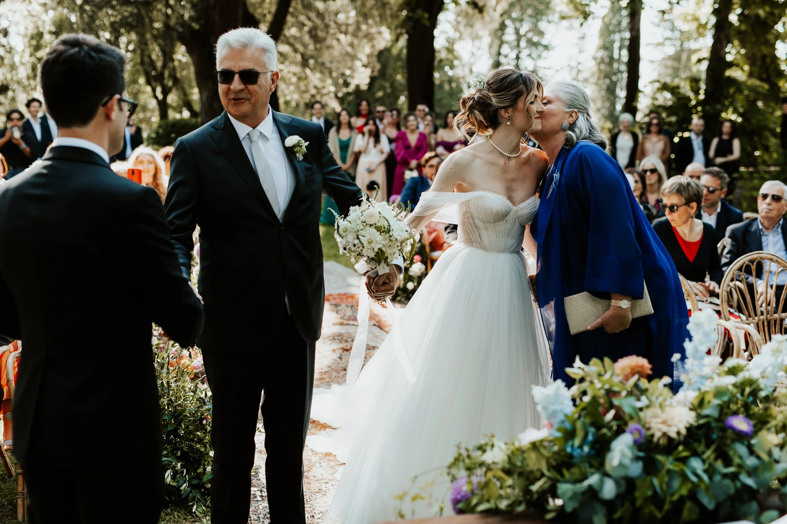 Una coppia di sposi si bacia durante il matrimonio all'aperto, circondata da amici e parenti seduti. La sposa indossa un abito bianco e tiene un bouquet, mentre la donna più anziana le dà un bacio sulla guancia. L'atmosfera è felice e romantica, con 