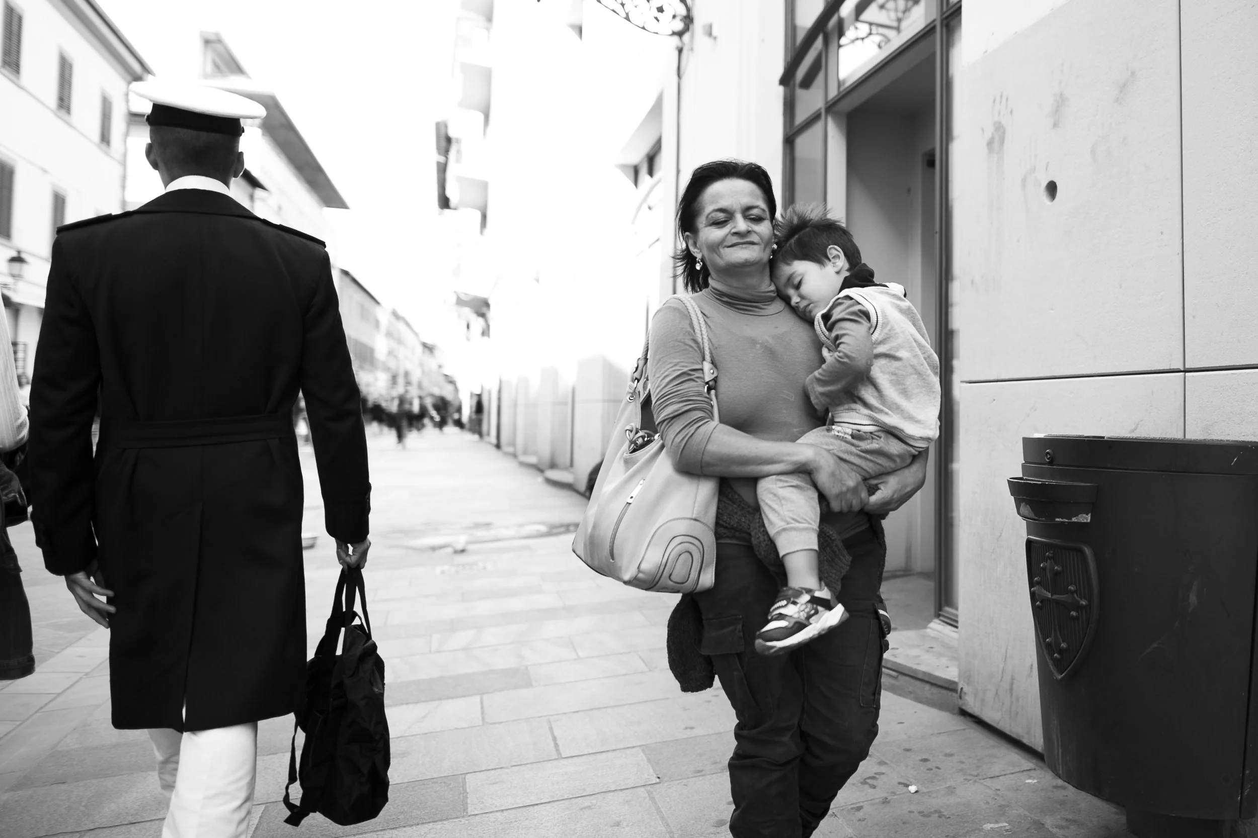 Una donna tiene un bambino appena uscito da un edificio in via urbana, camminando sulla strada. Un uomo con abiti da ufficiale militare o da polizia cammina in direzione opposta. Scene di strada in una città trafficata.