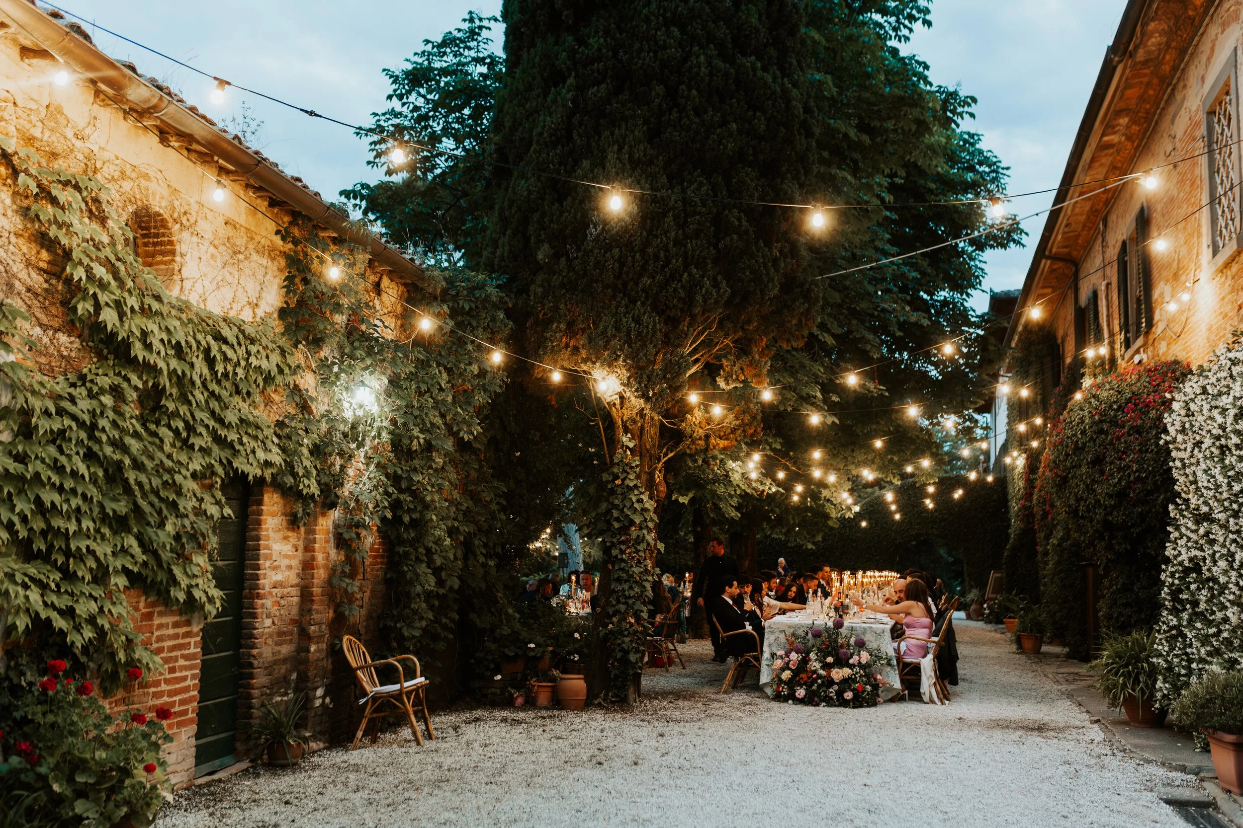 Cena all'aperto in un giardino sotto luci di stringa, con gruppi di persone sedute intorno a tavoli decorati con fiori, tra muri di mattoni ricoperti di edera e piante in vaso, durante la sera.