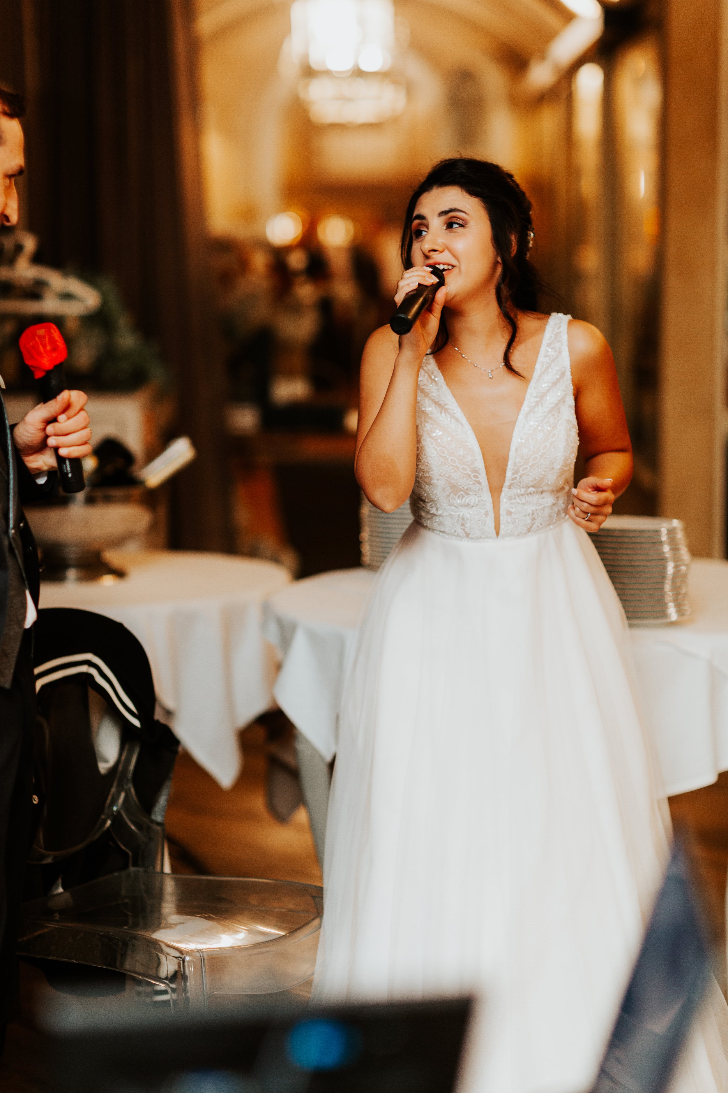 Una donna in abito da sposa che tiene un microfono durante un discorso o una canzone, in un'ambientazione di festa o matrimonio.
