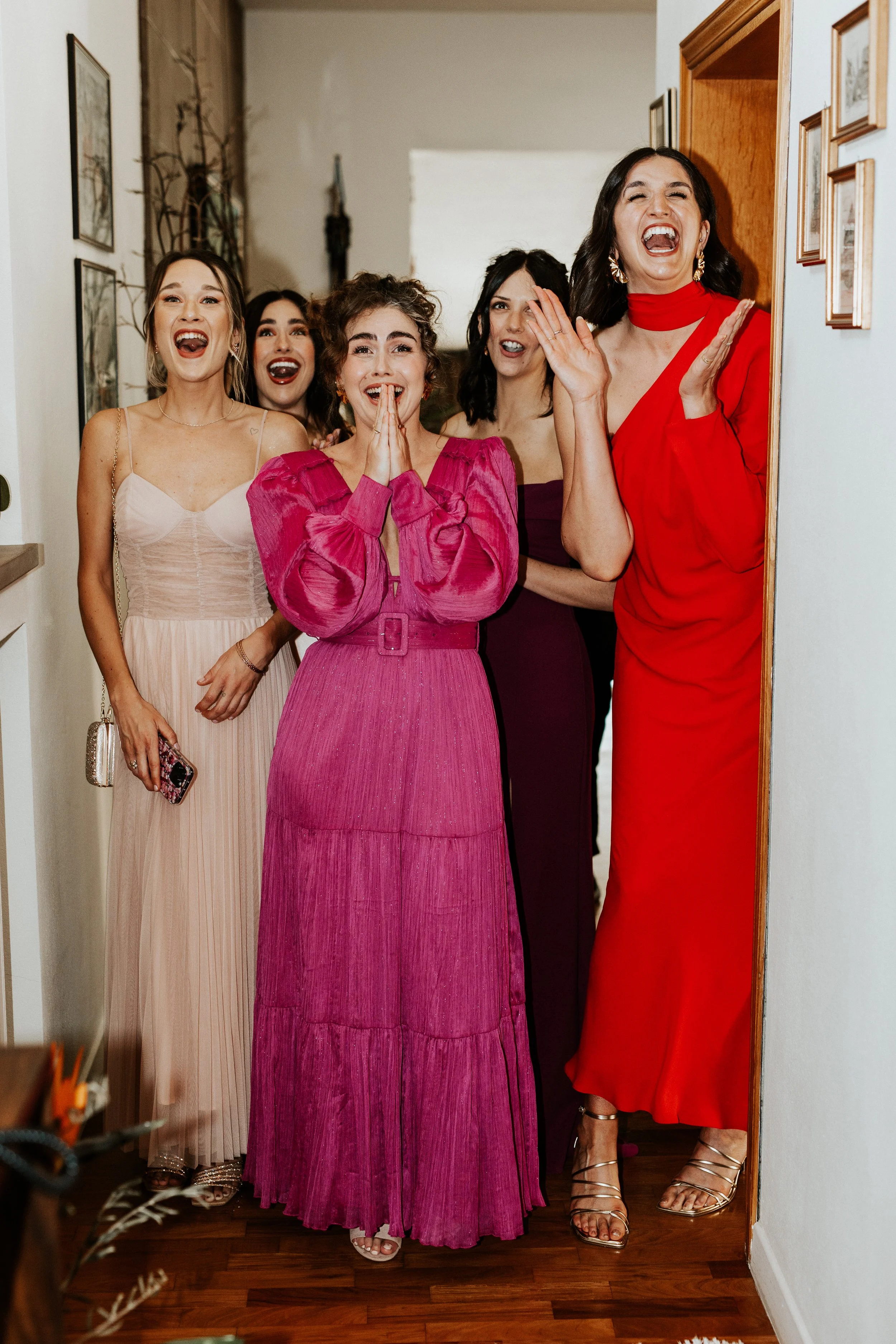 Un gruppo di sei donne sorridenti, eleganti e vestite con abiti lunghi colorati, che ridono e si divertono mentre si trovano in un corridoio domestico.