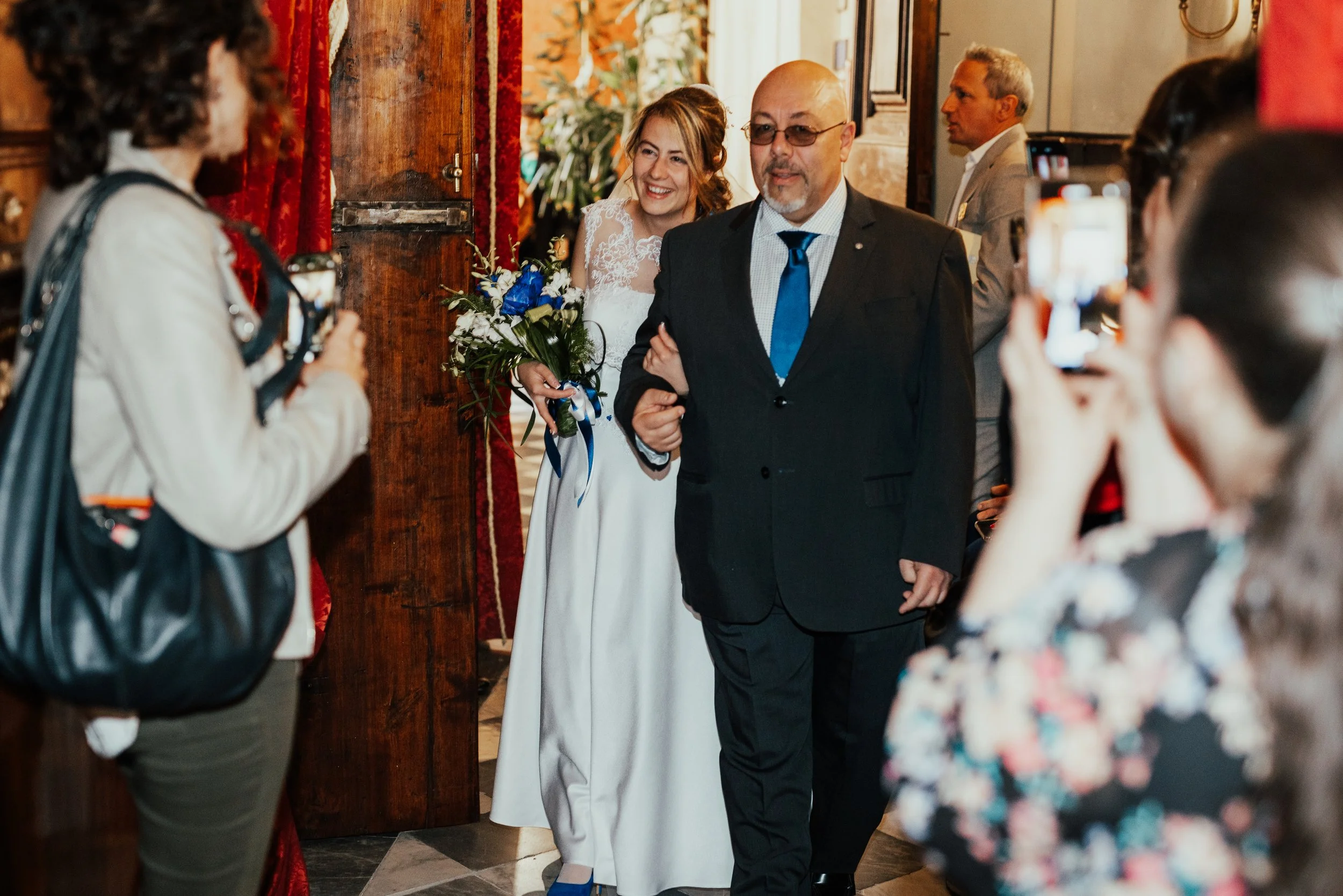 Sposa sorridente che tiene un mazzo di fiori e cammina con un uomo in un evento matrimonio, circondata da amici che scattano foto.