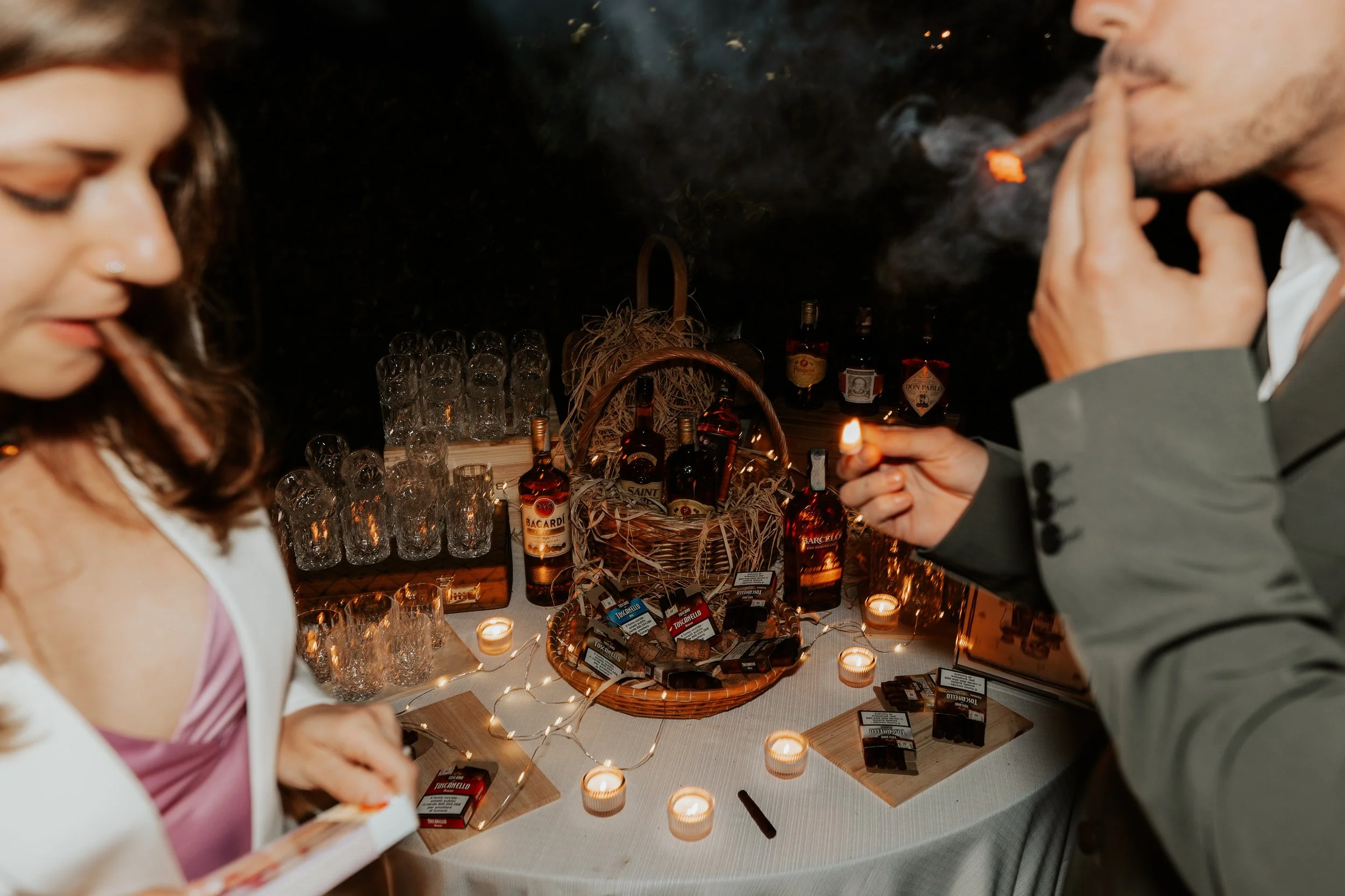 Due persone fumano sigarette in un'atmosfera tranquilla vicino a un tavolo decorato con candele e bottiglie di alcolici, con altri bicchieri e scatole di sigarette.
