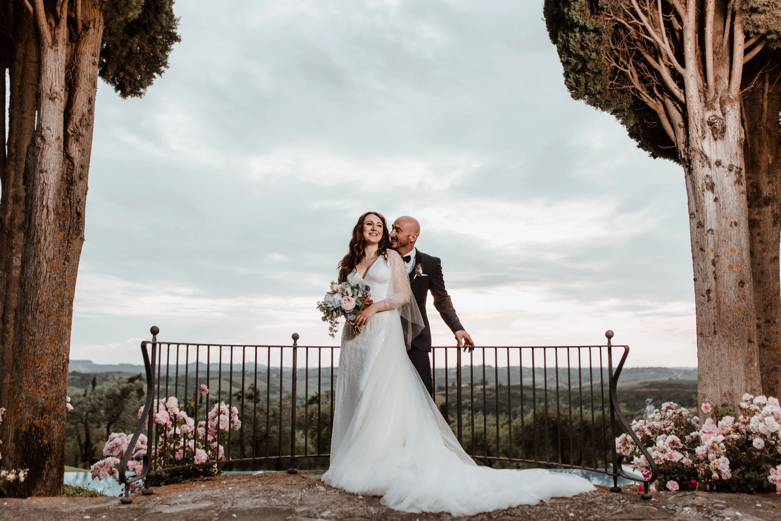 Una coppia di sposi in abiti da matrimonio, sorridenti e felici, si abbraccia davanti a un paesaggio naturale con alberi e un cielo nuvoloso, circondati da fiori rosa e una ringhiera nera, in un’ambientazione all’aperto.