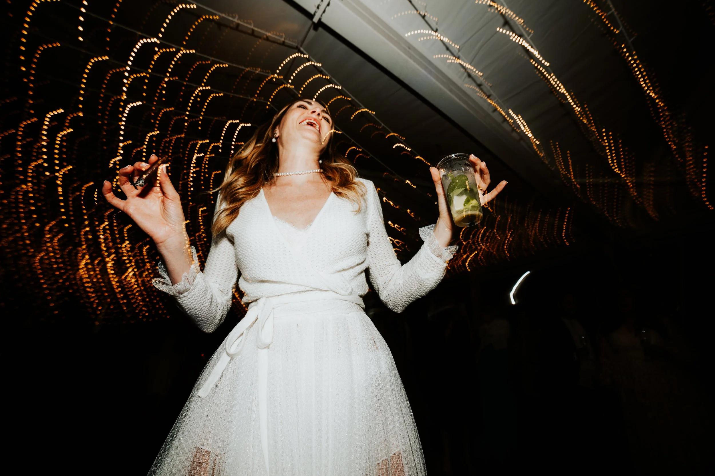 Una donna sorridente con abito bianco elegante che balla in un ambiente con luci calde e sottile decorazione luminosa, mentre tiene un bicchiere di mojito con lime e menta.