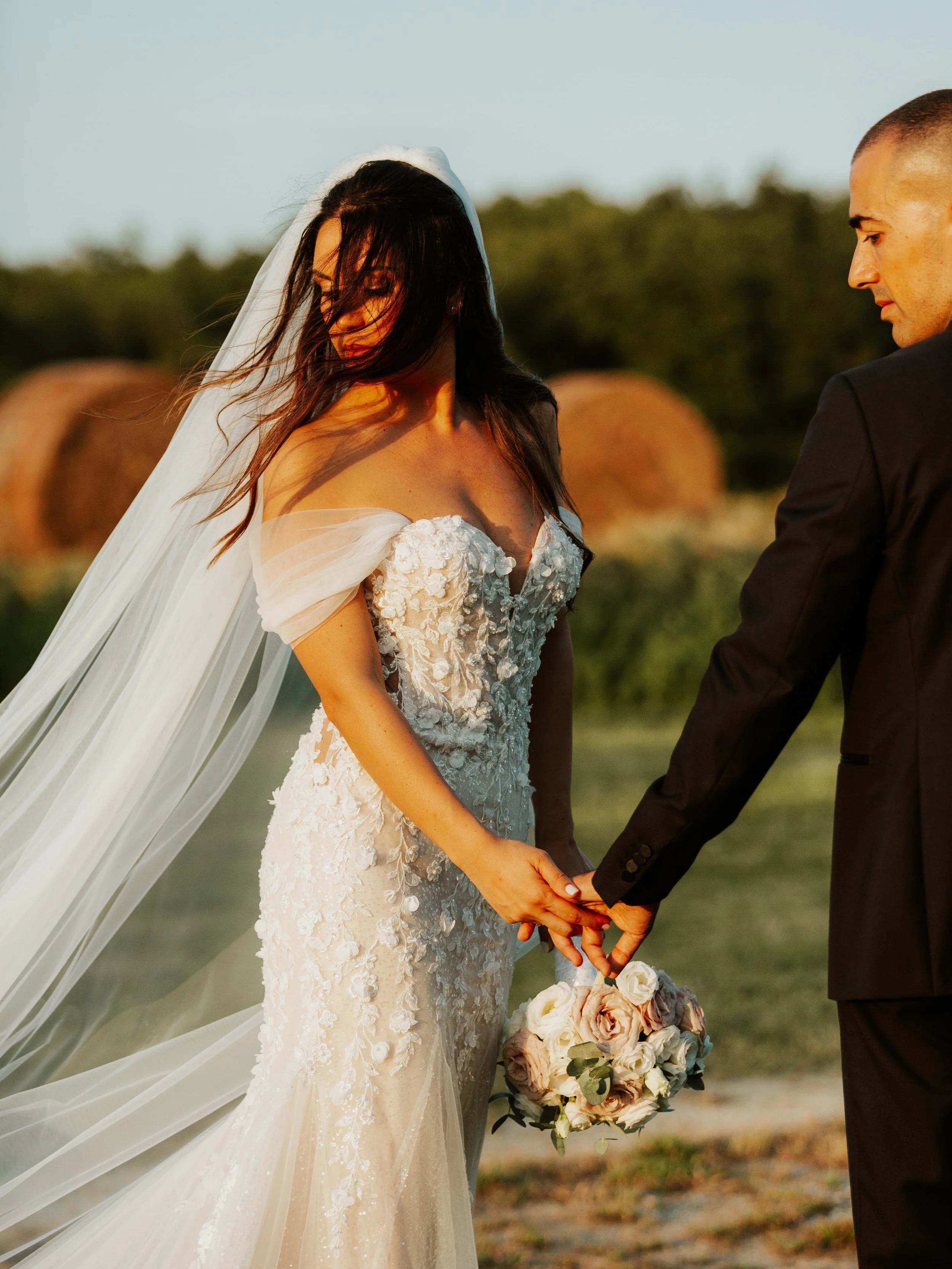 Cerimonia di nozze con una sposa in abito bianco e uno sposo in giacca nera che si tengono per mano, con un bouquet di fiori, in un ambiente all'aperto con campagna e balle di fieno sullo sfondo.