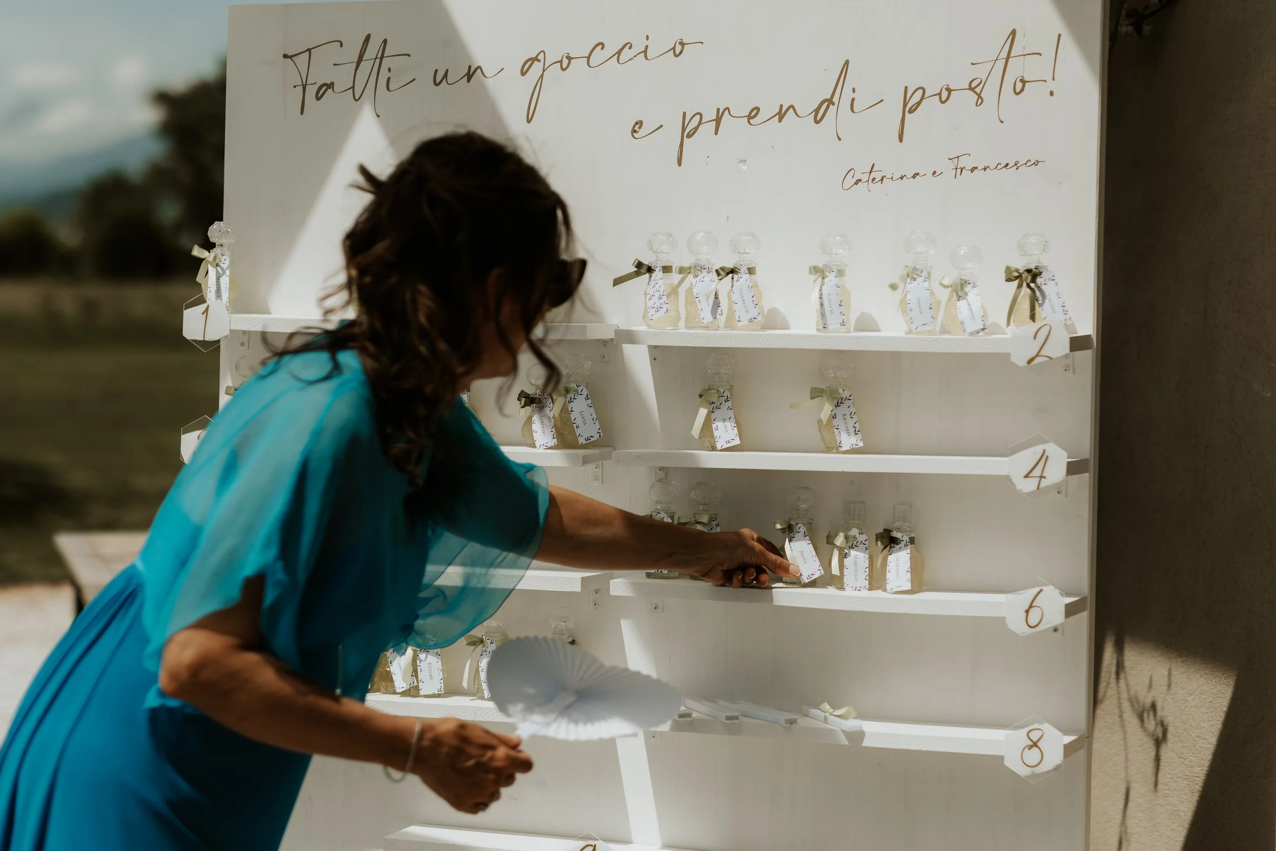 Una donna con vestito turchese che pone un biglietto su una bottiglia su uno scaffale bianco decorato con etichette e nastri, sotto un cartello scritto in italiano 'Fatti un ricordo e prenditi posto! Caterina e Francesco' in un ambiente all'aperto.