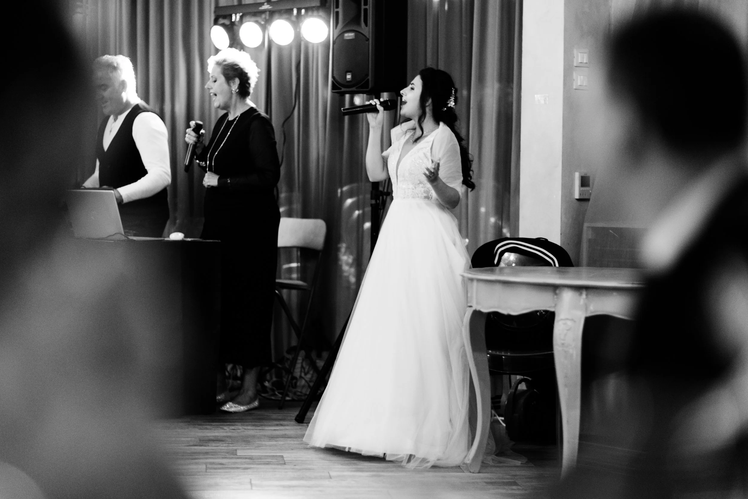 Una donna in abito da sposa canta con un microfono, accompagnata da altre persone sul palco in un ambiente intimo.