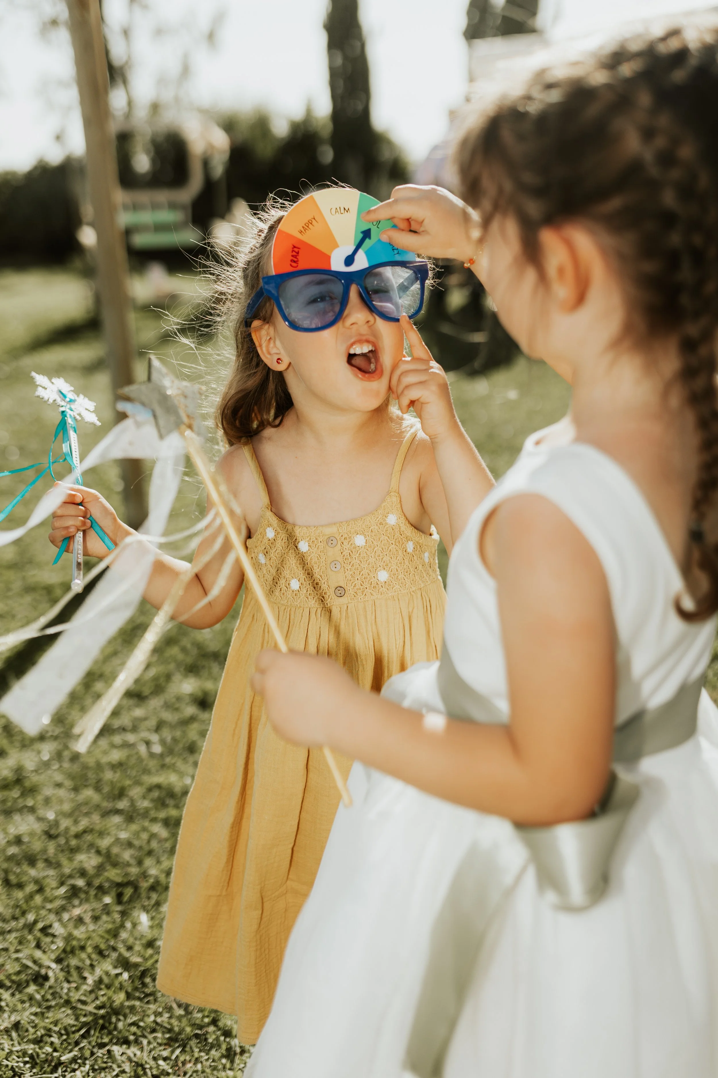 Due bambine che giocano all'aperto, una con un cappello decorato con un indicatore di emozioni e l'altra con un abito bianco, mentre una le misura o sistema il cappello.