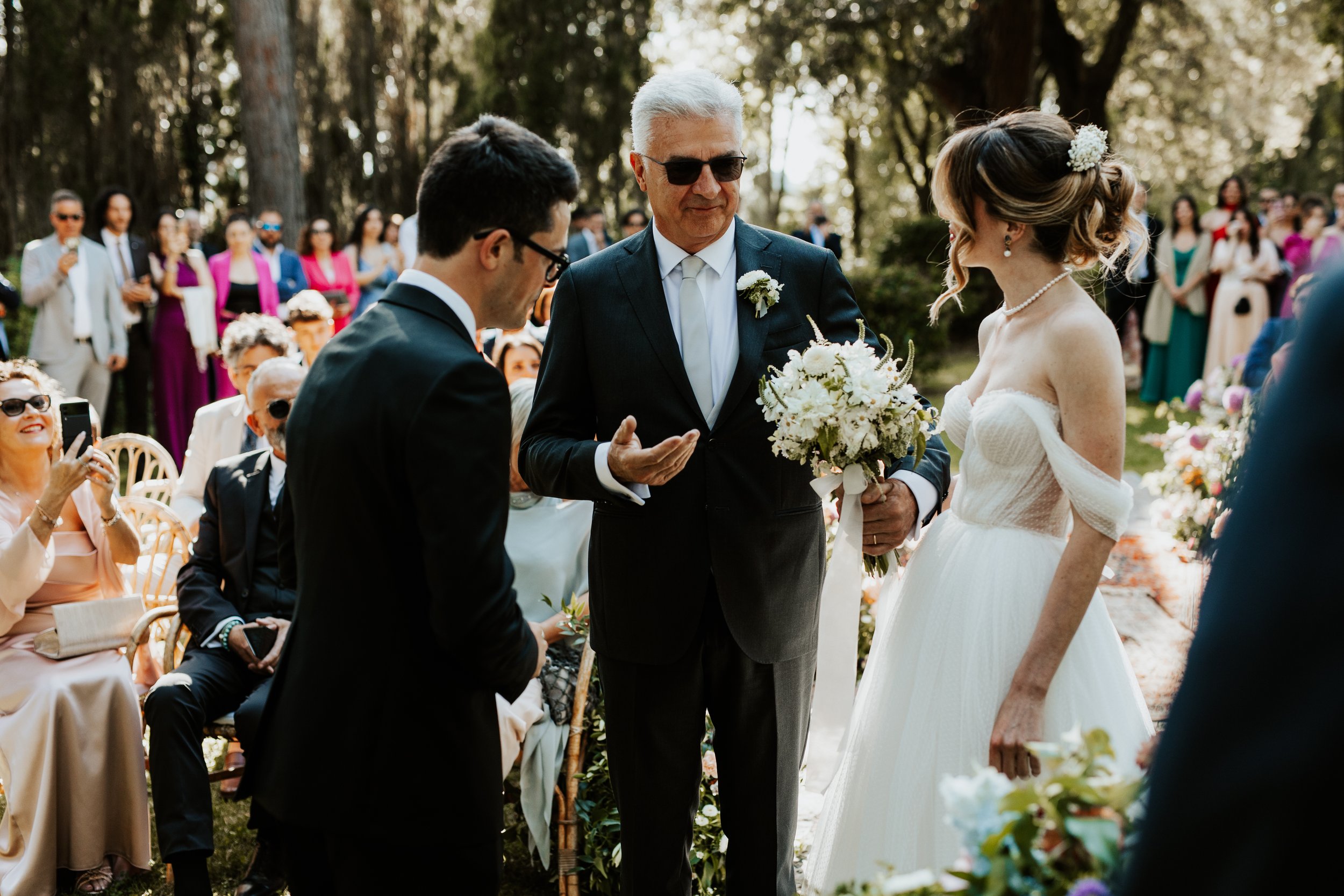Una scena di matrimonio all'aperto con il matrimonio, uno sposo, una sposa e un celebrante, circondati da invitati seduti e in piedi, in un ambiente naturale con alberi in background.
