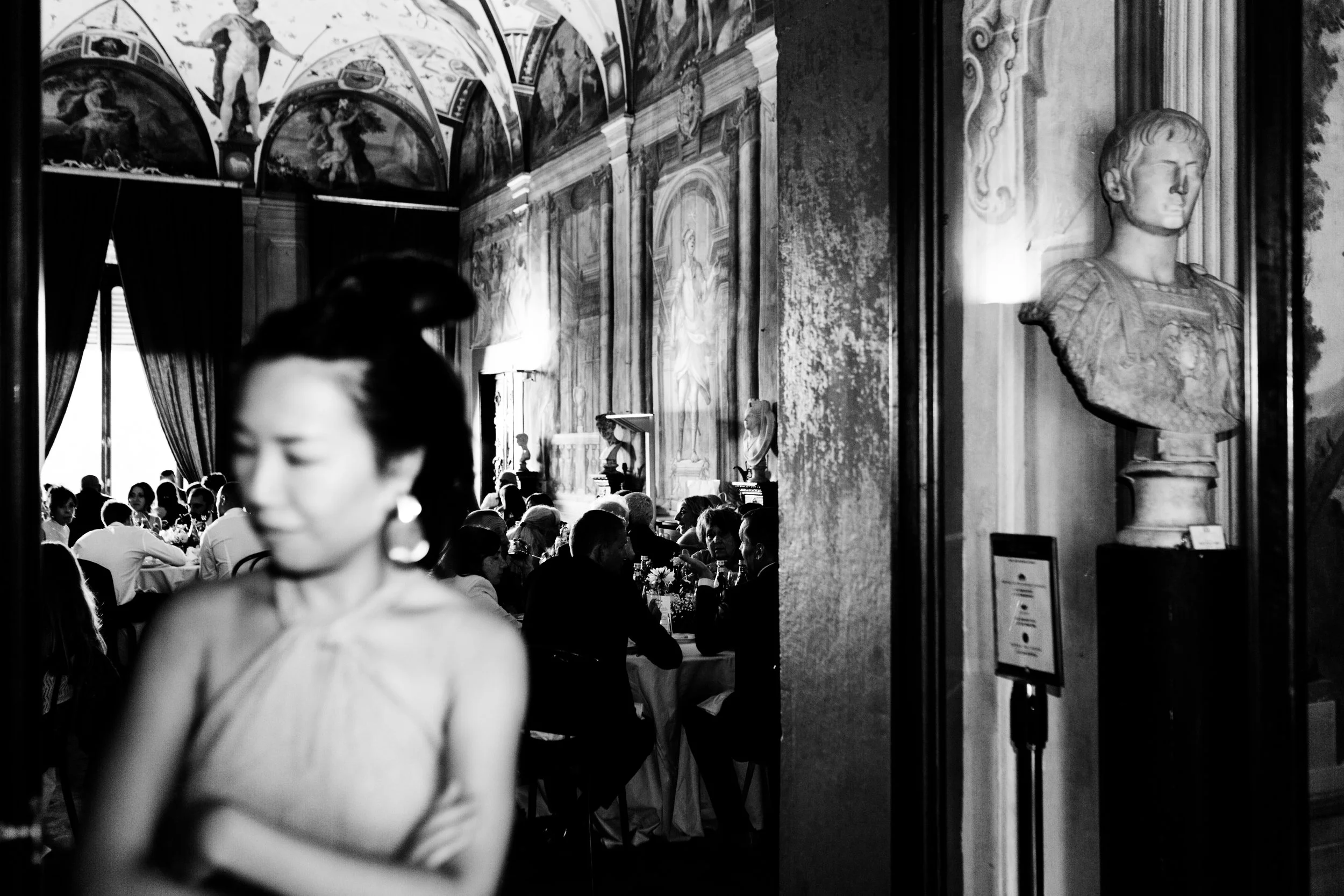 Una sala in un museo o un ristorante elegante con pareti decorate da affreschi e statue. In primo piano, una donna con abito elegante e capelli raccolti, sfocata. Sullo sfondo, persone sedute a tavoli che socializzano e mangiano, con grandi finestre 