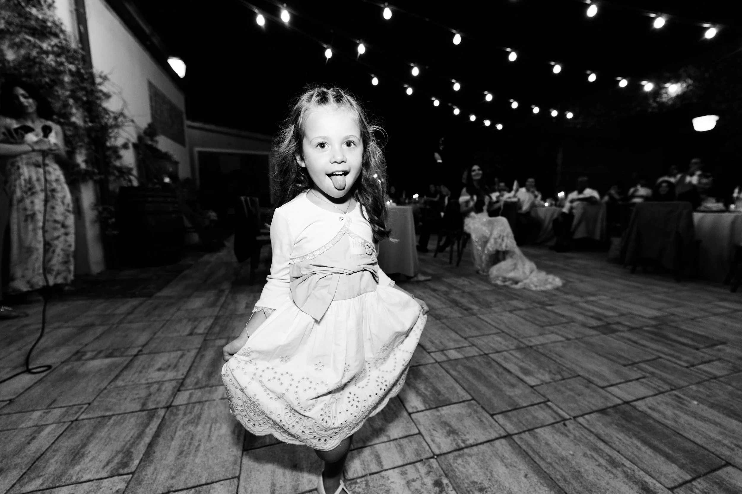 Bambina sorridente che gioca e fa il gesto della lingua all'aperto di notte, con tavoli e persone sullo sfondo, decorazioni luminose.