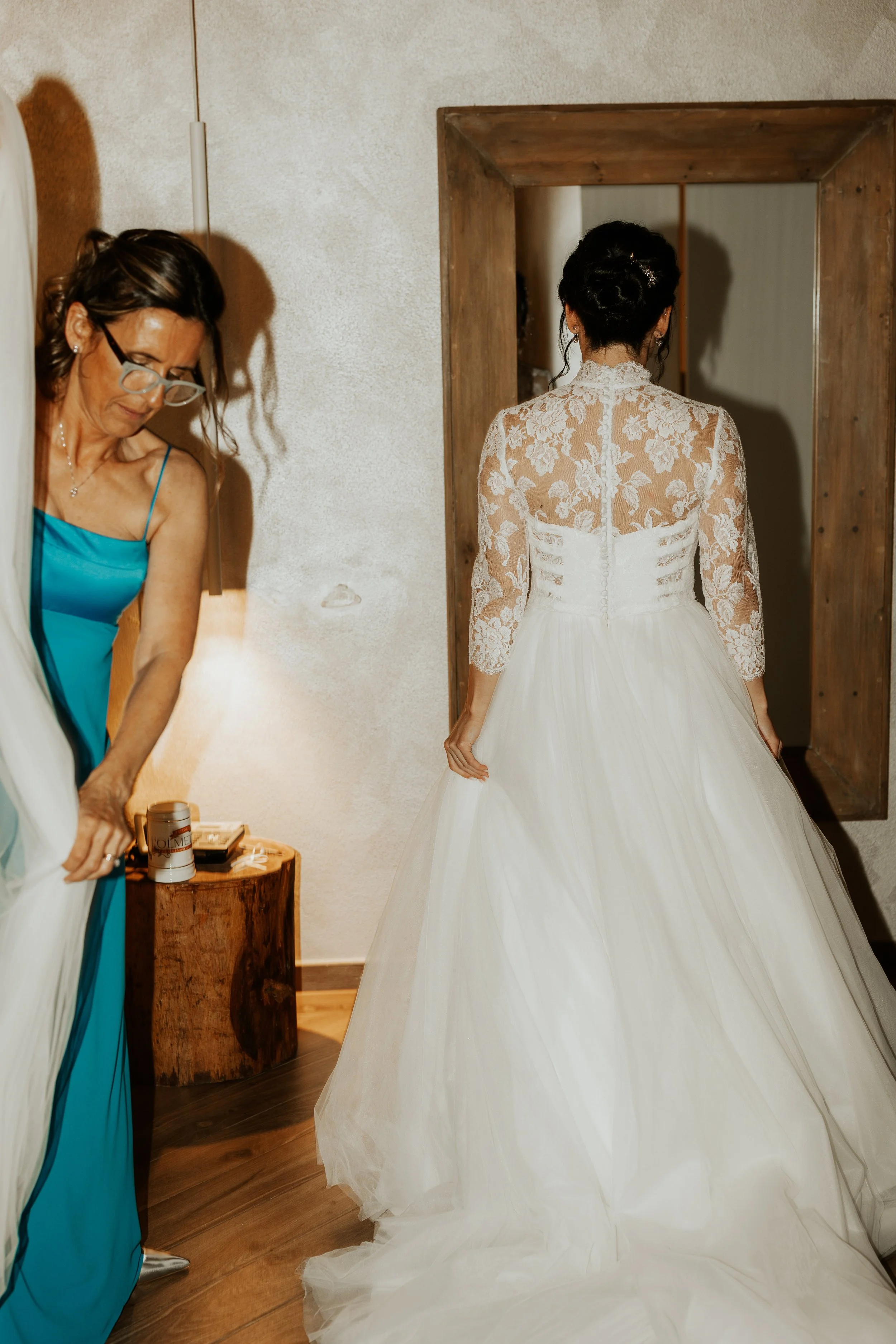Una sposa con un abito bianco e cuciture di pizzo sulla schiena, visto di schiena, mentre si prepara per il matrimonio in una stanza accogliente con pareti di pietra e un'area in legno.