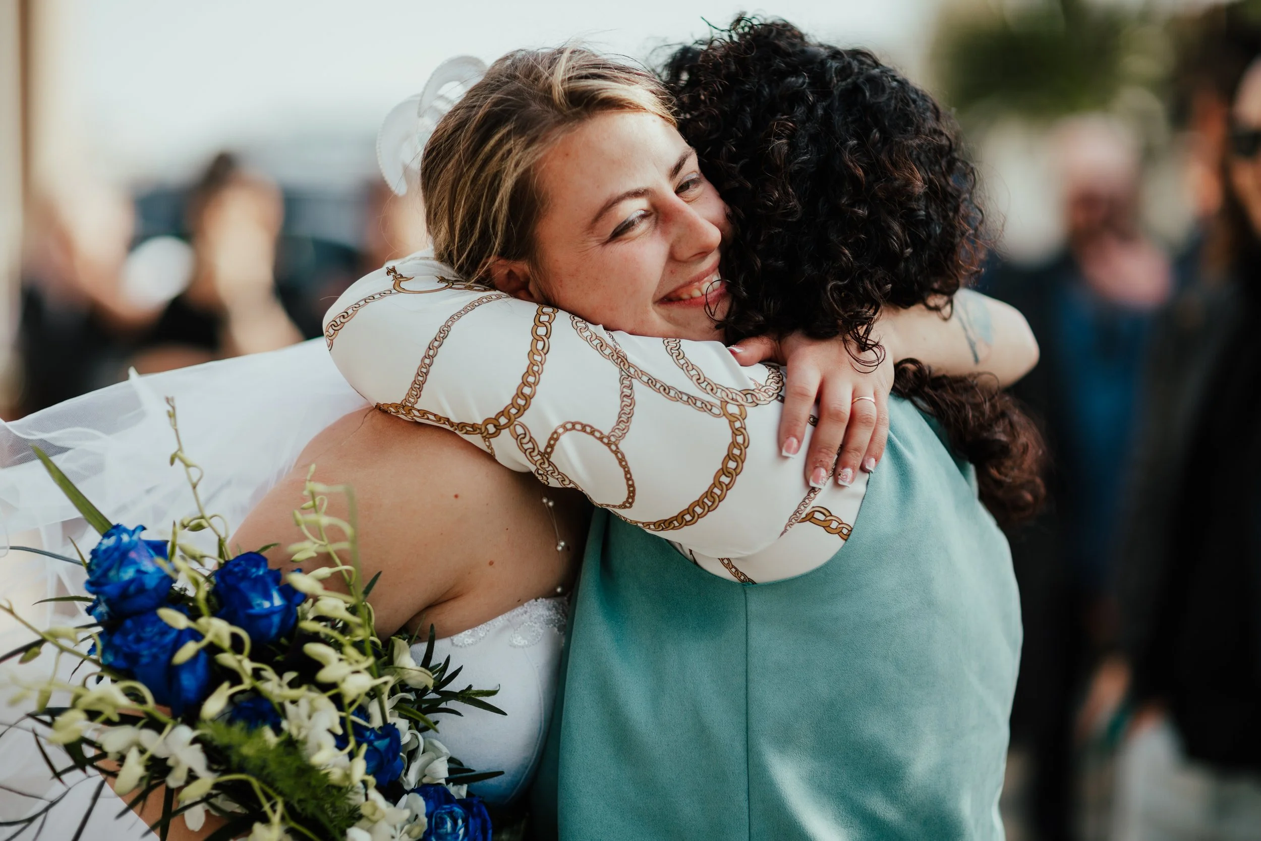 Due donne si abbracciano felicemente, probabilmente a un matrimonio, con uno sfondo di persone sorridenti. Una donna indossa un abito bianco e tiene un bouquet di fiori blu.