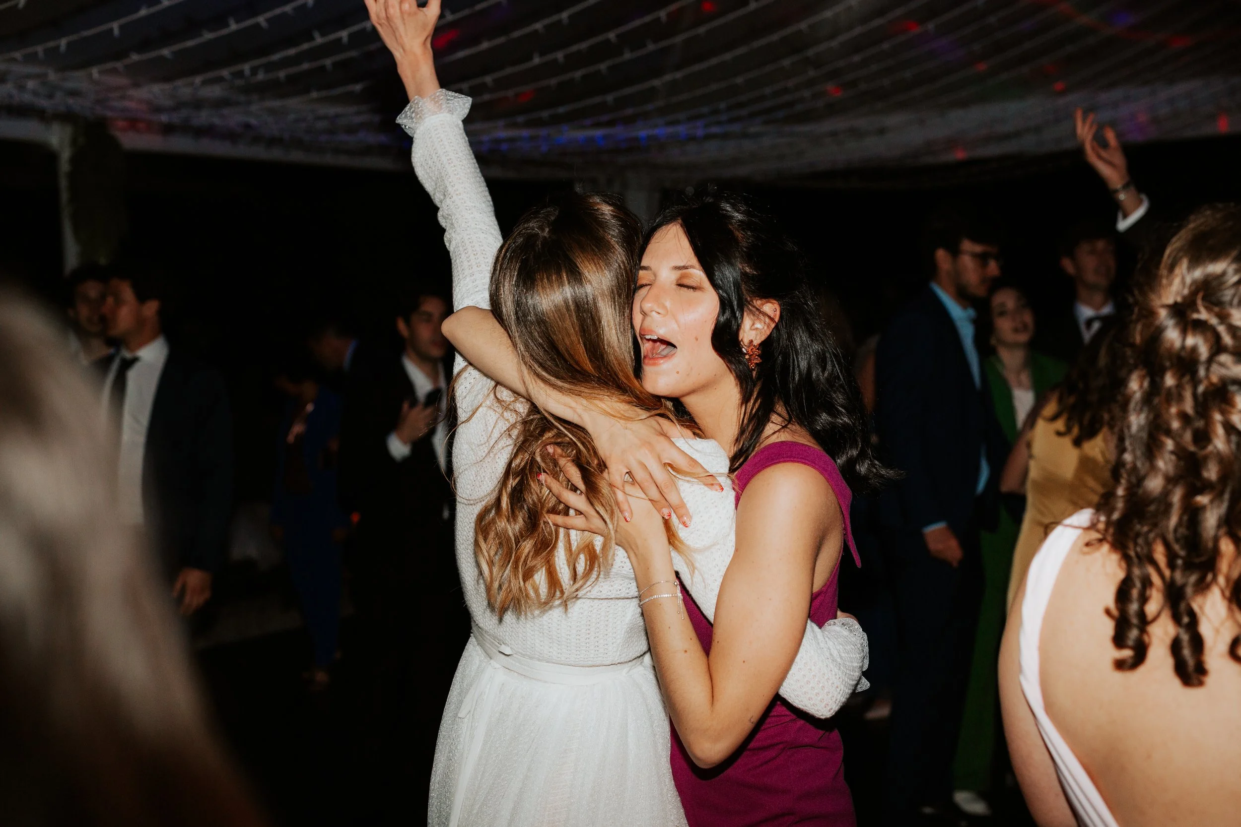Due donne abbracciate e felici mentre ballano a una festa o matrimonio in un ambiente affollato, con altre persone sullo sfondo.