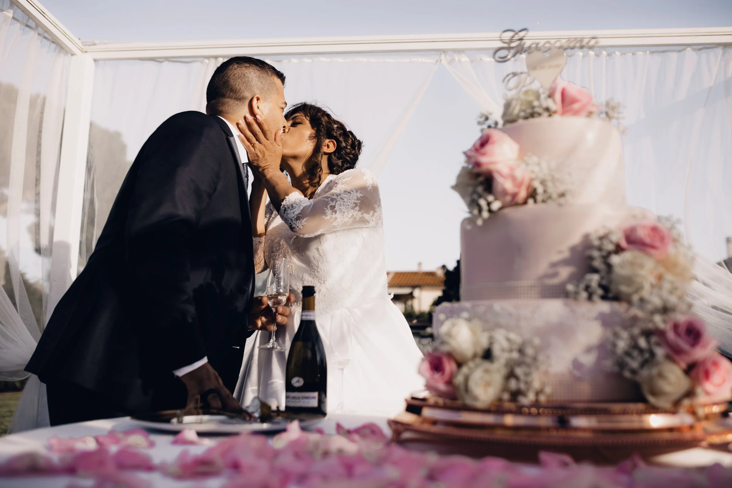 Coppia di sposi che si bacia durante il matrimonio, con torta nuziale decorata con fiori rosa e bianchi sul tavolo.