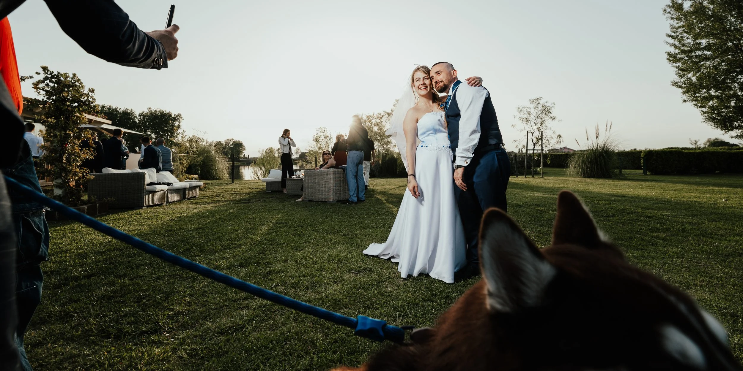 Fotografia di matrimonio con sposi che sorridono, fotografie di altri invitati sullo sfondo, animali domestici nell'area all'aperto, scena di festa all'aperto al tramonto.