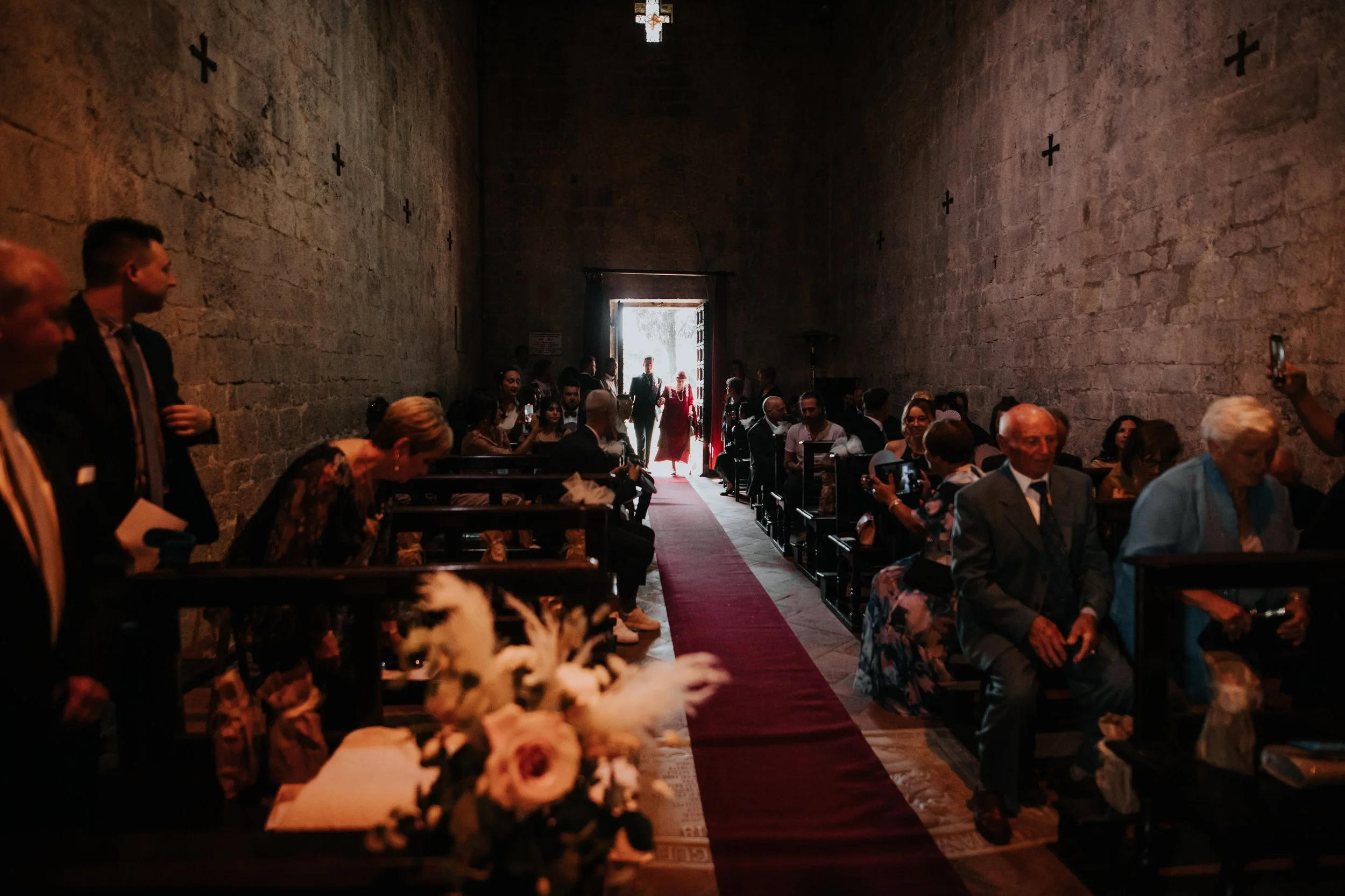 Interior di una chiesa con persone sedute in attesa di un matrimonio, con un tappeto rosso che porta all'entrata illuminata dalla luce del sole.