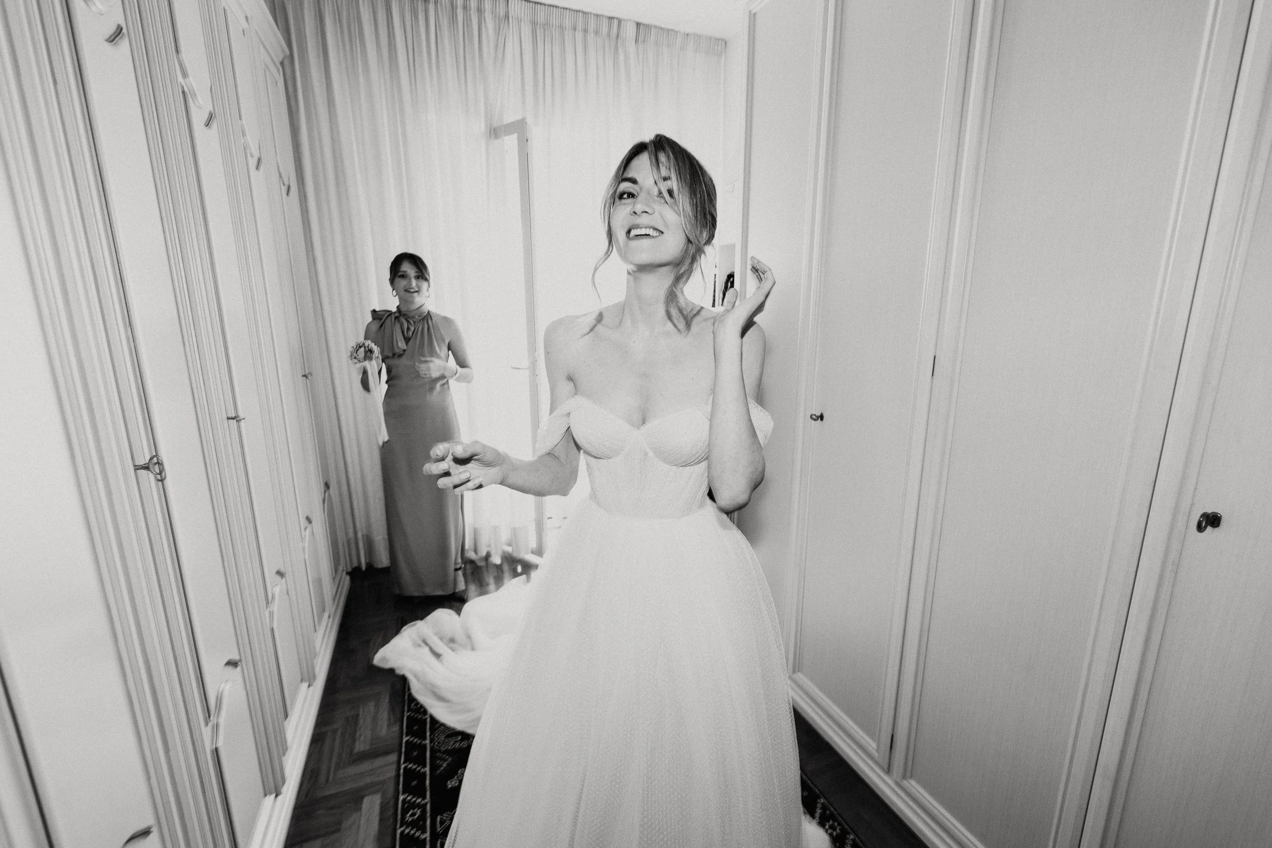 Donna in abito da sposa che sorride e fa un gesto con la mano, con un'altra donna in abito lungo che tiene un mazzo di fiori sullo sfondo, in una stanza con armadi su entrambi i lati.