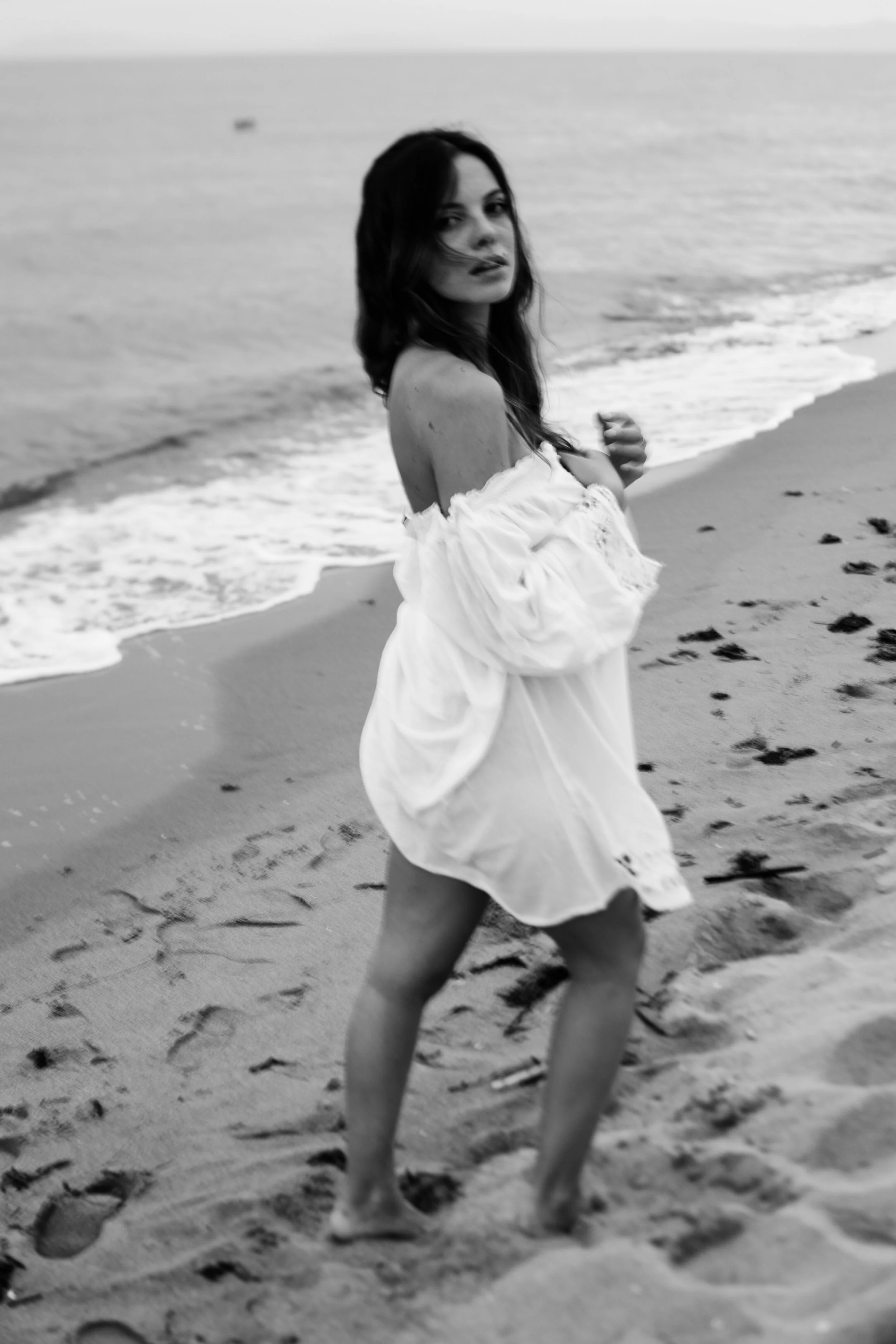 Donna con vestito bianco sulla spiaggia, con mare sullo sfondo, in bianco e nero.