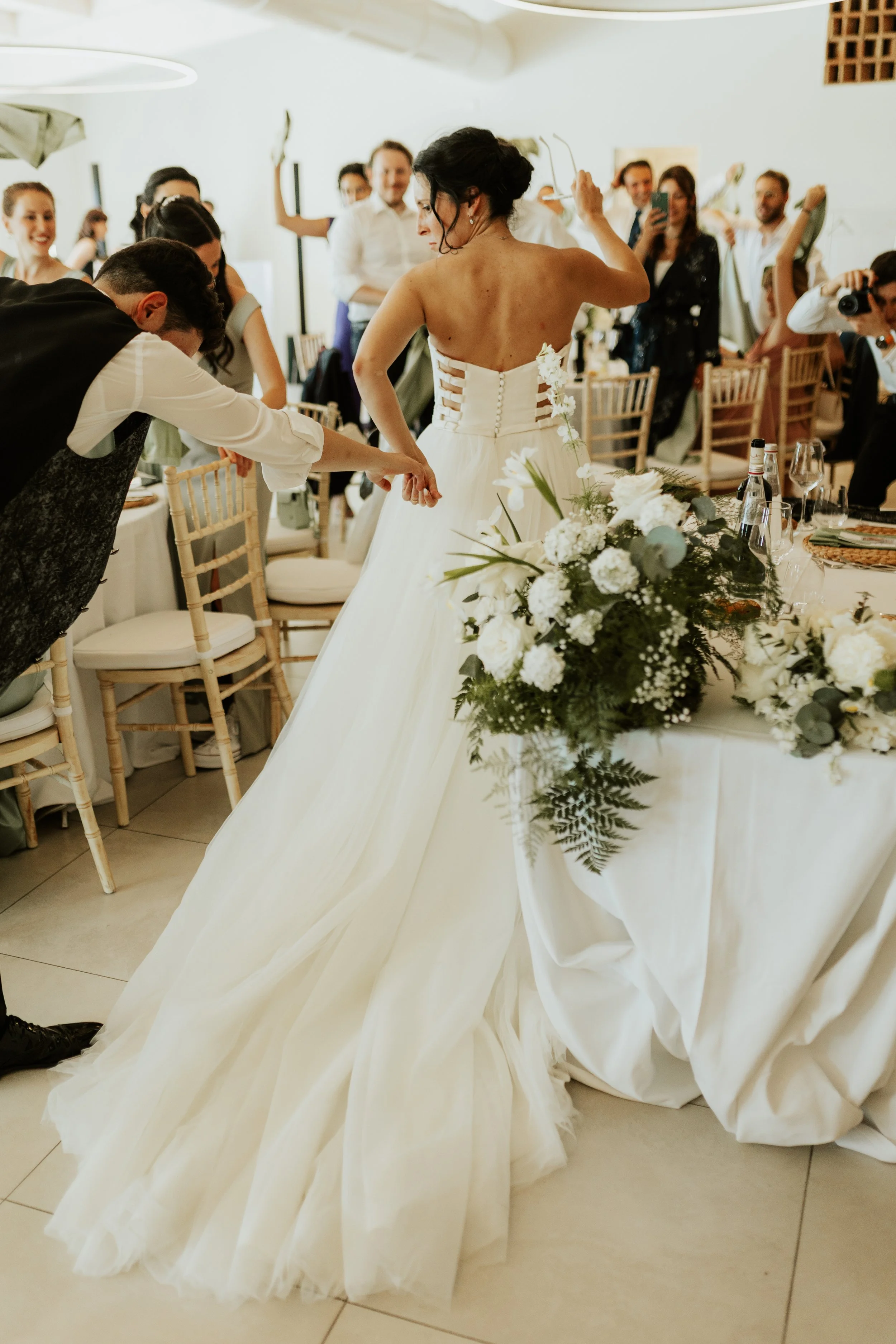 Una donna in abito da sposa in un ricevimento nuziale, circondata da amici e famiglia, con molti fiori bianchi e decorazioni eleganti.