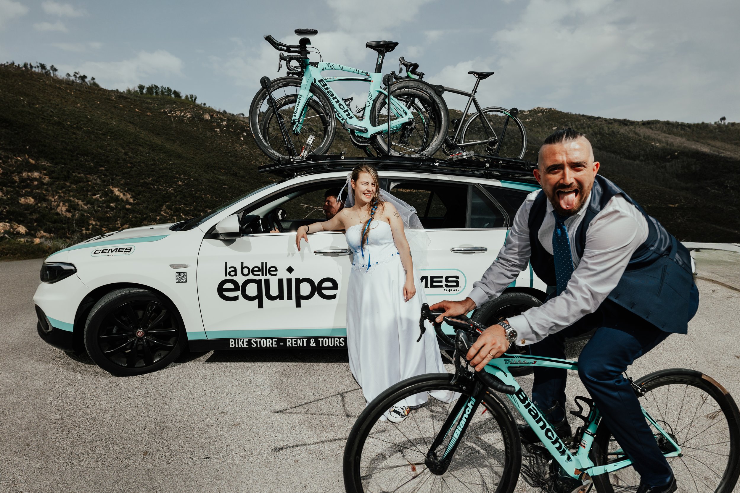 Un uomo sorridente con camicia e giacca sulla bicicletta, una donna in abito bianco che si appoggia a una macchina bianca con le scritte "la belle equipe", una persona seduta al volante, tutte con biciclette e biciclette da corsa sul tetto, in un pae
