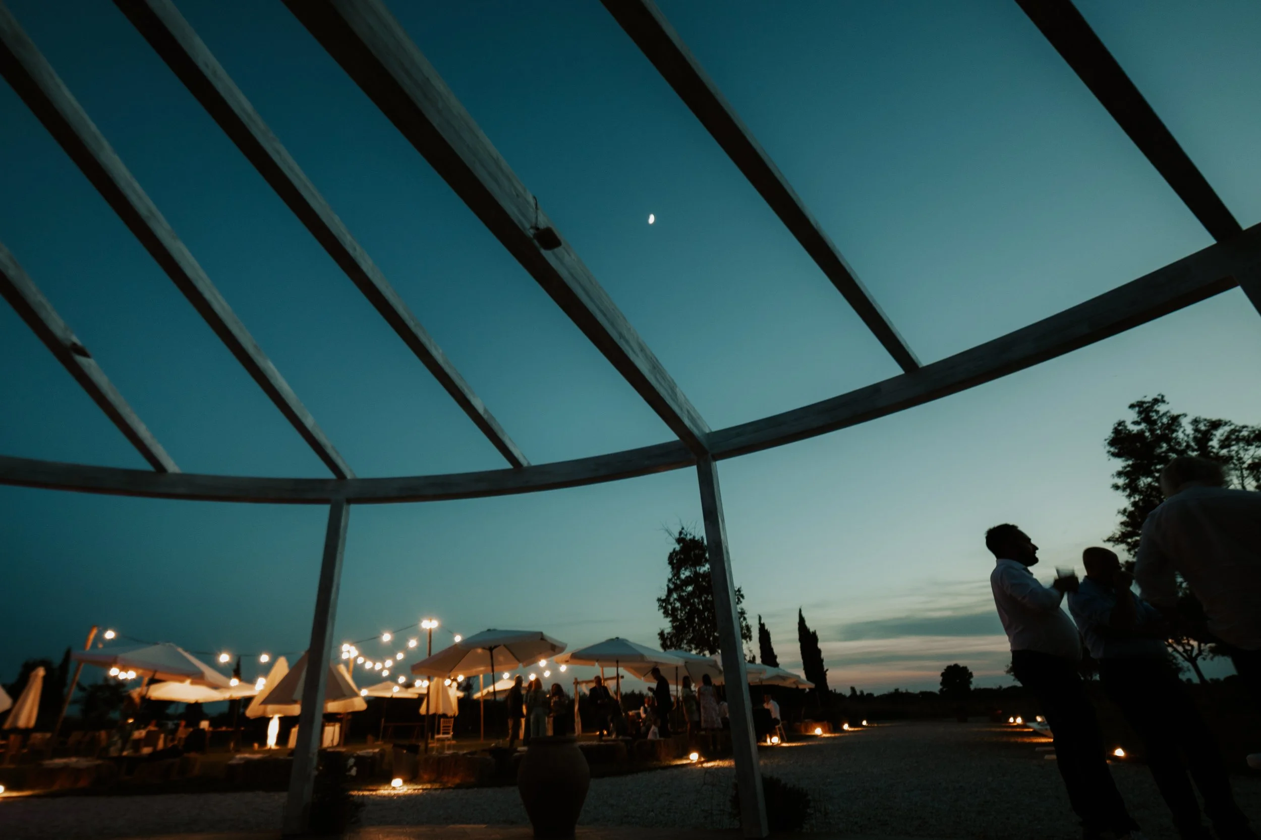 Serata in un giardino con persone che chiacchierano sotto candele e ombrelloni, vista al tramonto con cielo blu e luna