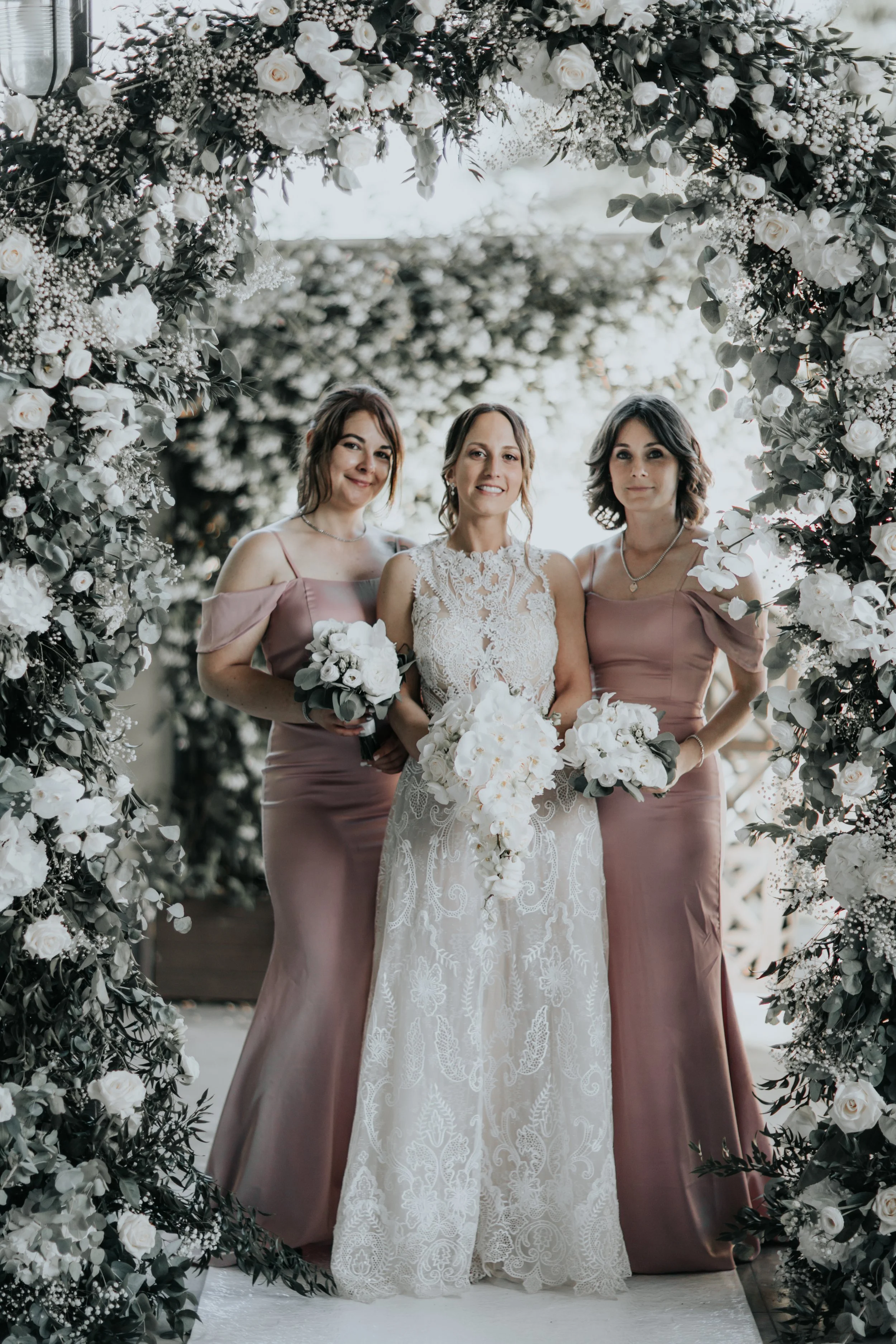 Tre donne in abiti da sposa e damigella, circondate da un arco di fiori bianchi e verdi, durante un matrimonio.