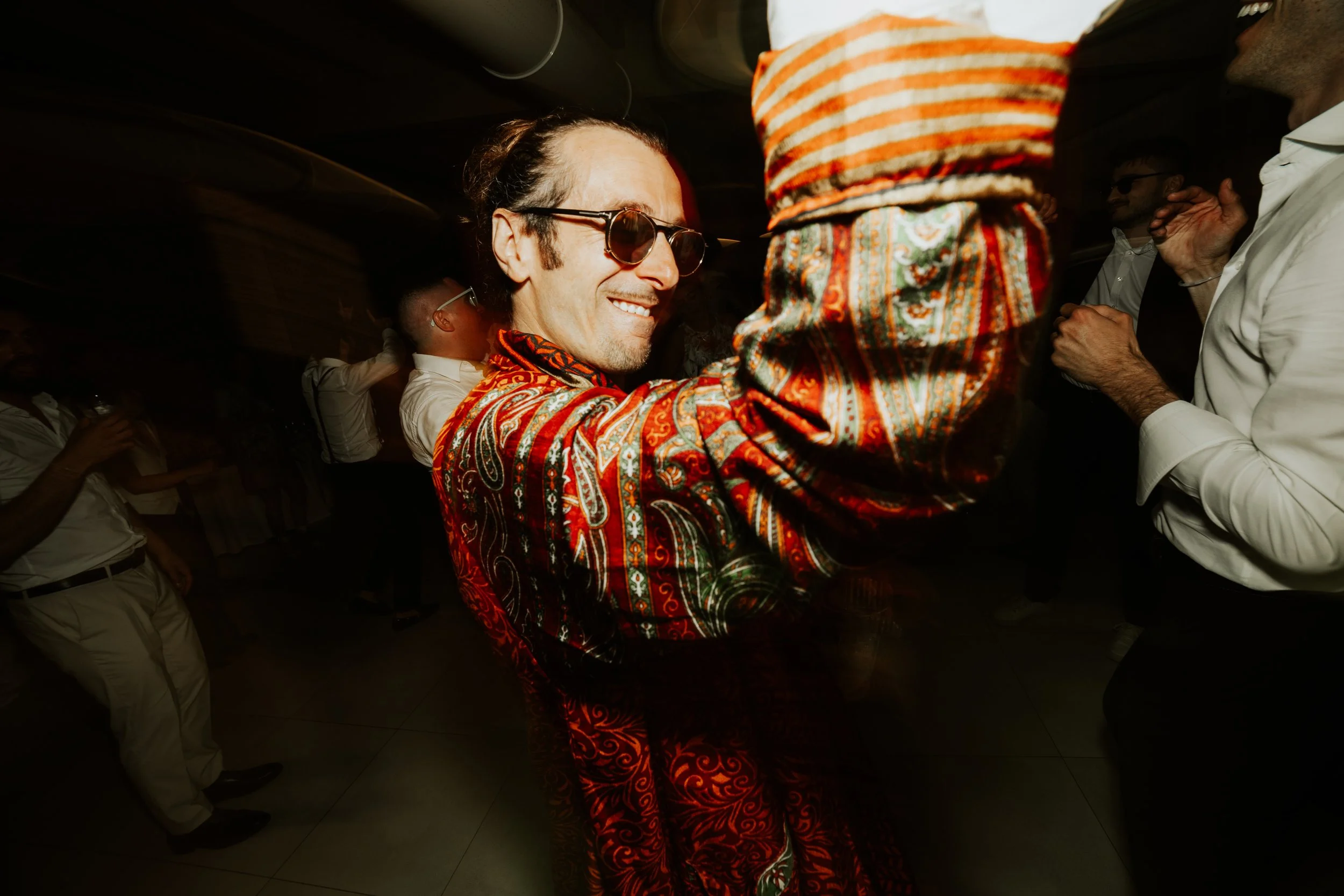 Uomo sorridente con occhiali da sole e camicia decorata che balla in una festa, circondato da altre persone che si divertono.