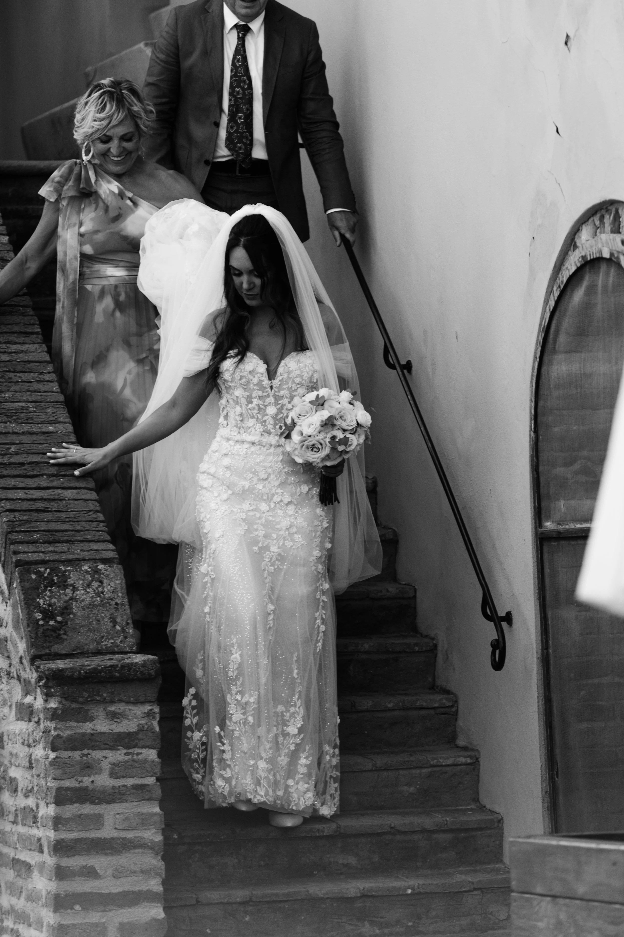 Una sposa con abito da matrimonio e velo scende una scala di pietra con accompagnatori, un uomo e una donna, in un ambiente interno, in bianco e nero.