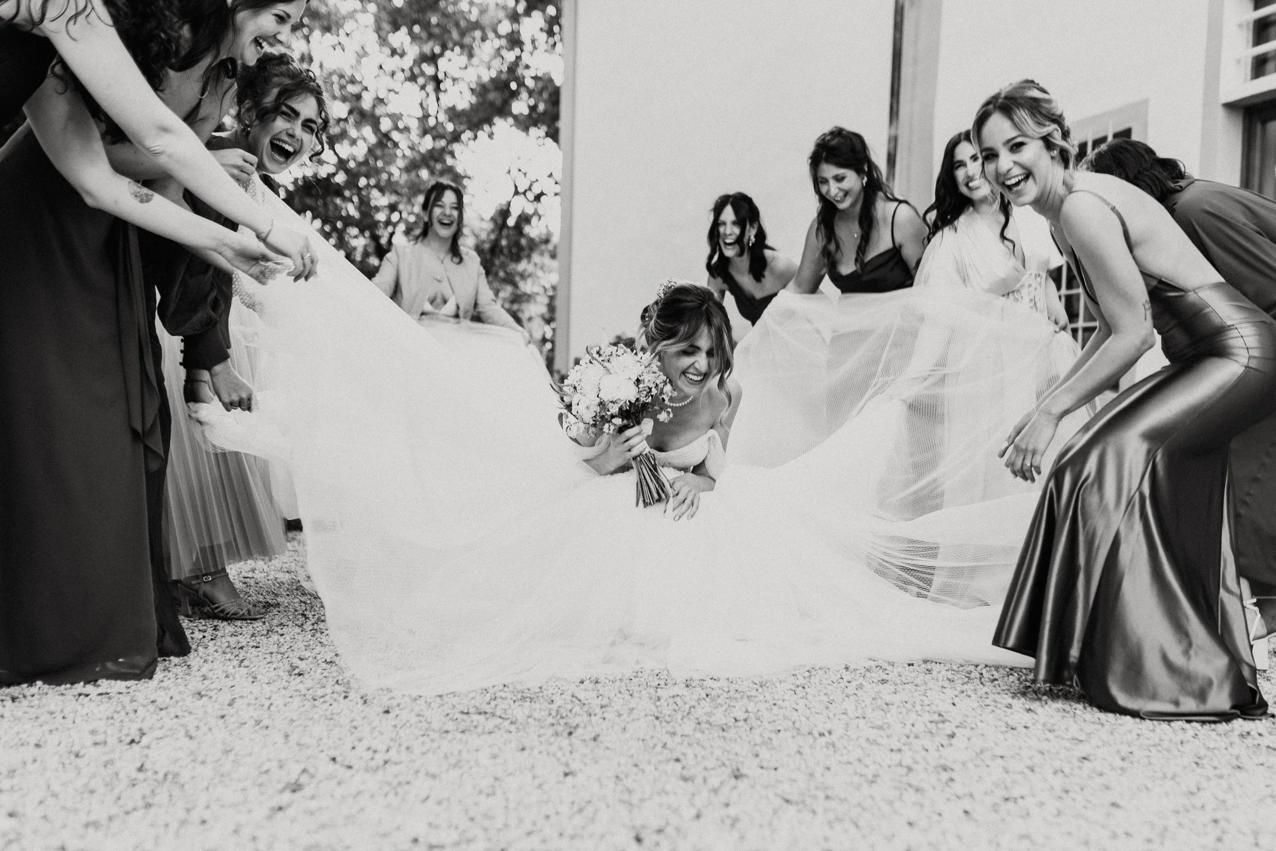 Foto in bianco e nero di una sposa con un bouquet di fiori e diverse amiche che la circondano ridendo e giocando con il velo del suo abito da sposa.