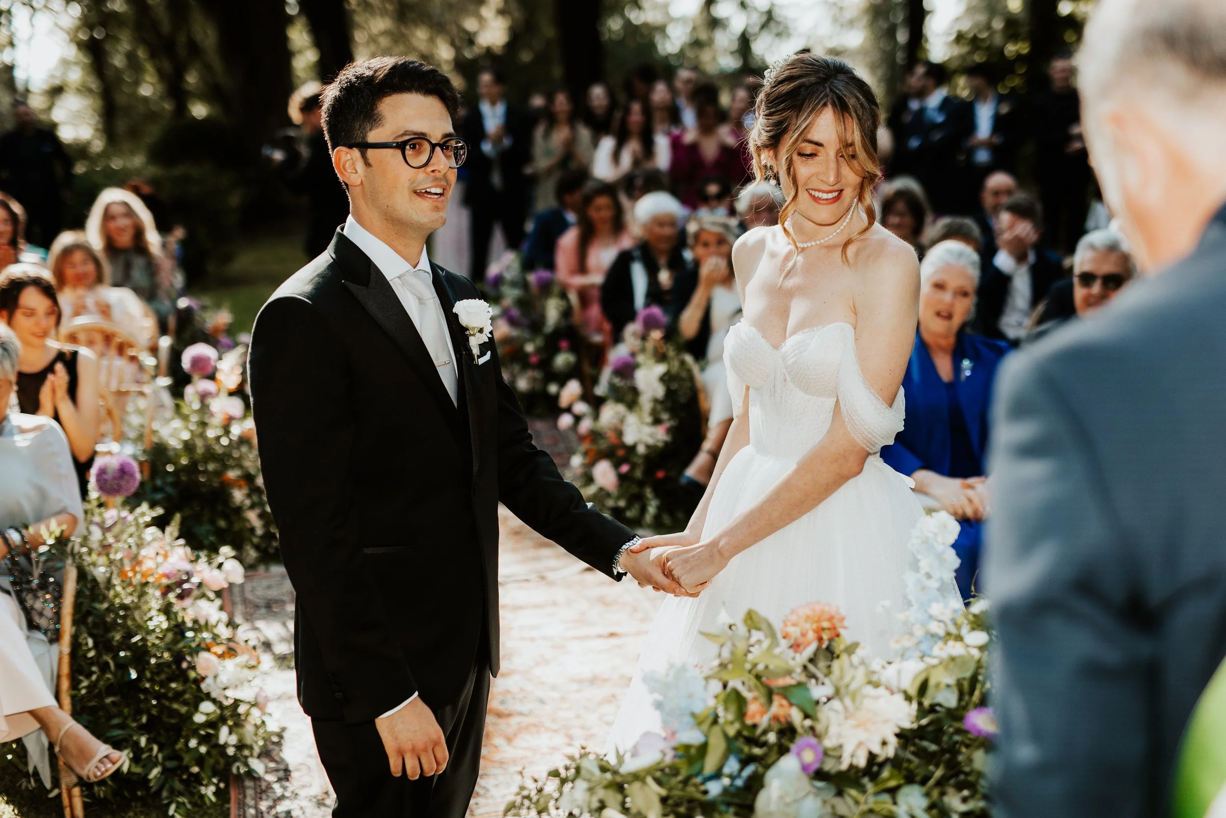 Una coppia di sposi si tiene per mano durante il matrimonio all'aperto, circondati da amici e familiari, con decorazioni floreali.