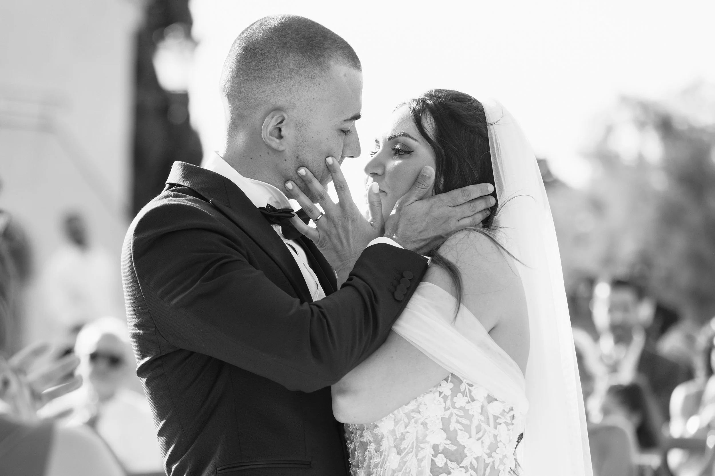 Una coppia di sposi si bacia durante il matrimonio, con il cielo e alcuni ospiti sullo sfondo.