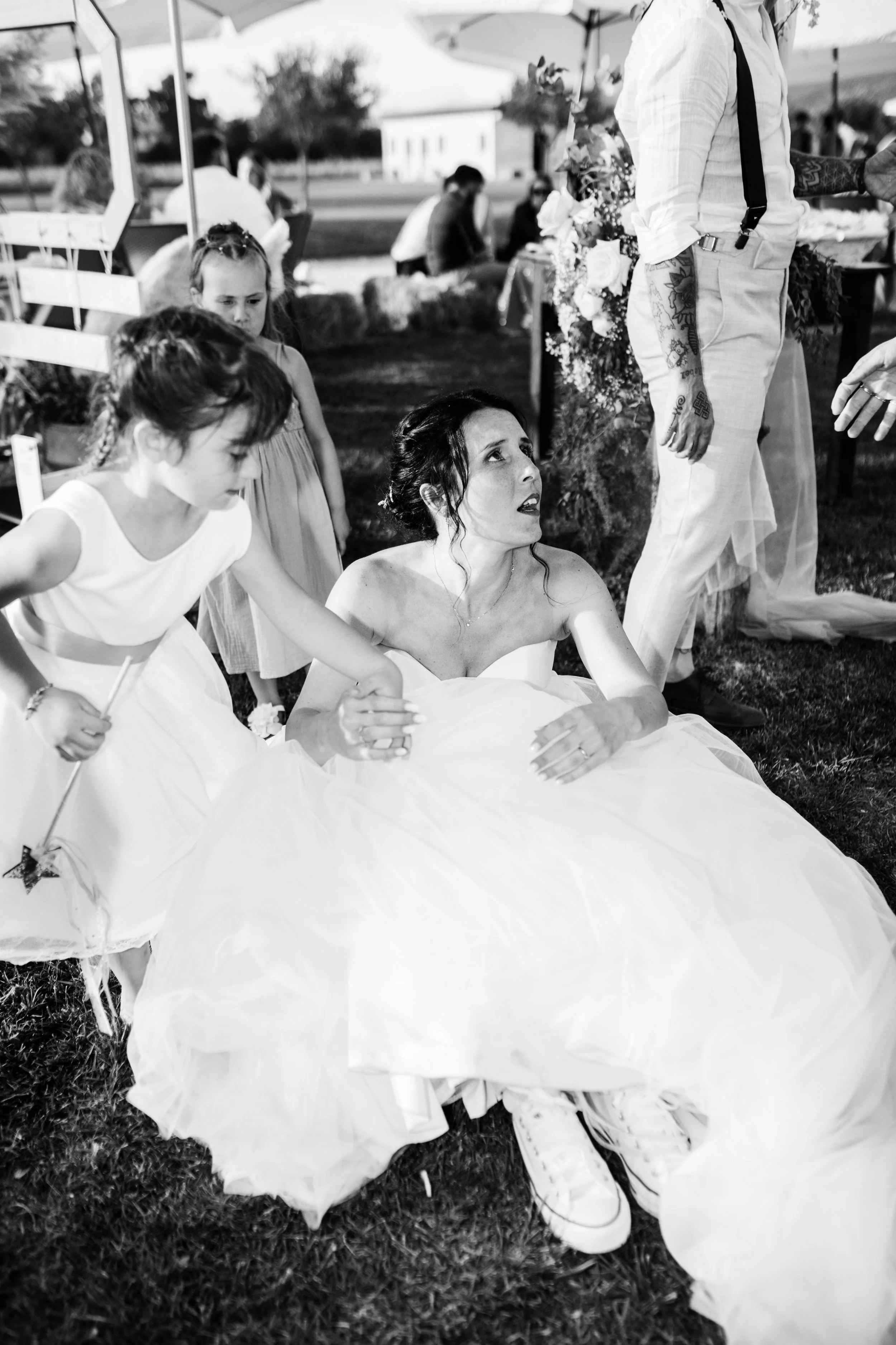 Donna in abito da sposa seduta sul prato, circondata da bambini e altre persone durante un matrimonio all'aperto.